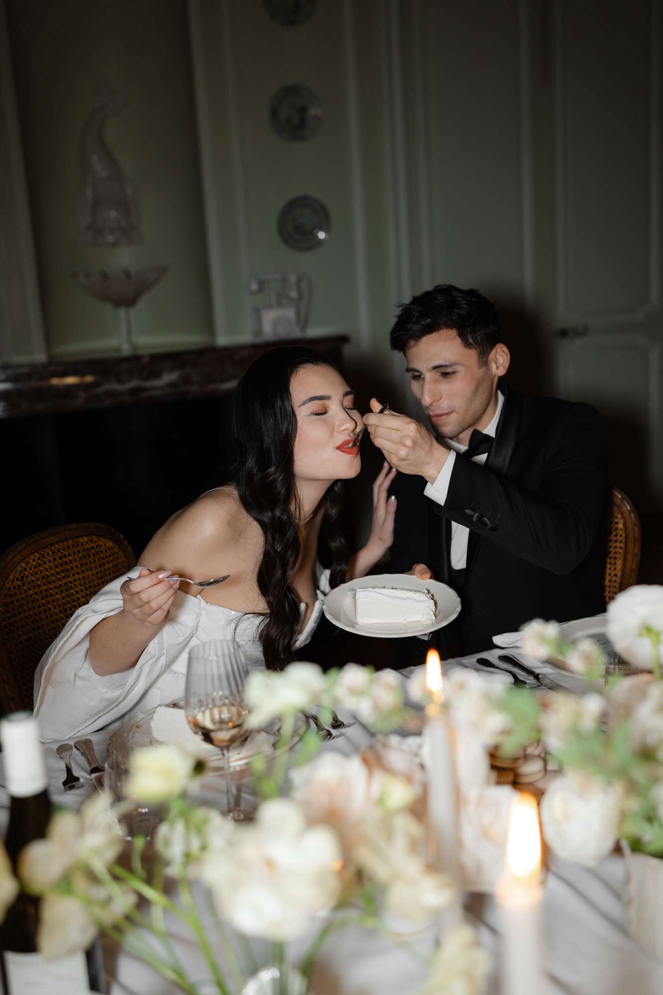 Groom feeds cake to bride at dining table in elegant pale green classical room during reception dinner