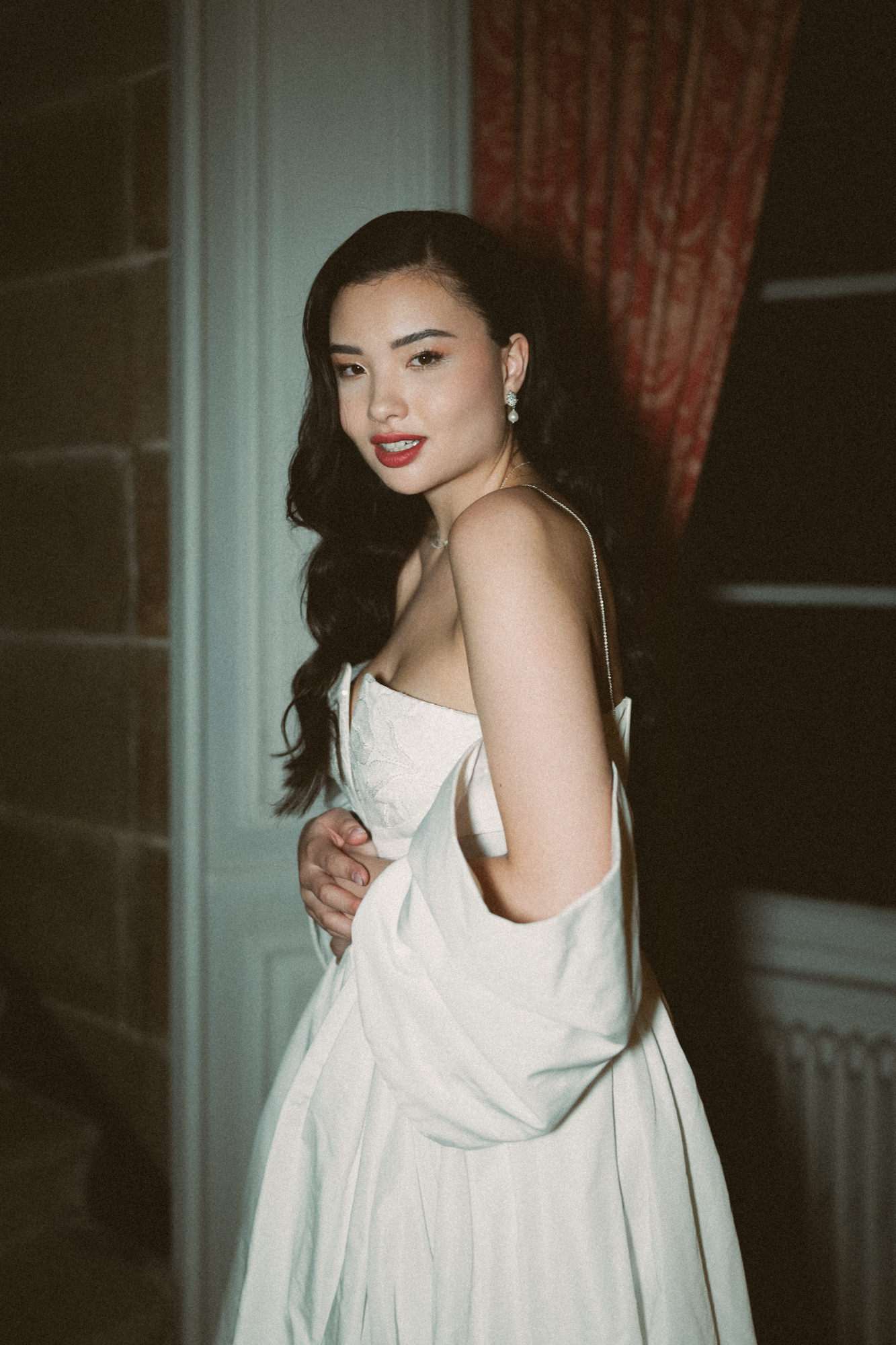 Bride looking over shoulder in ivory slip dress with pearl earrings and red lipstick against blue-green wall