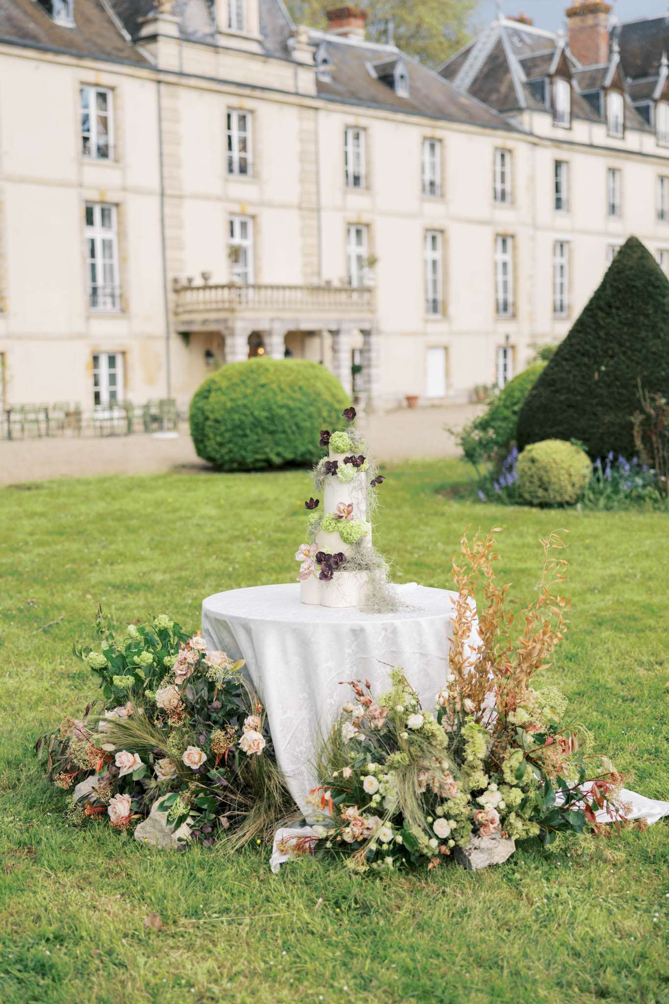Multi-tier white cake with burgundy hellebores and blush roses amid botanical ground florals before chateau