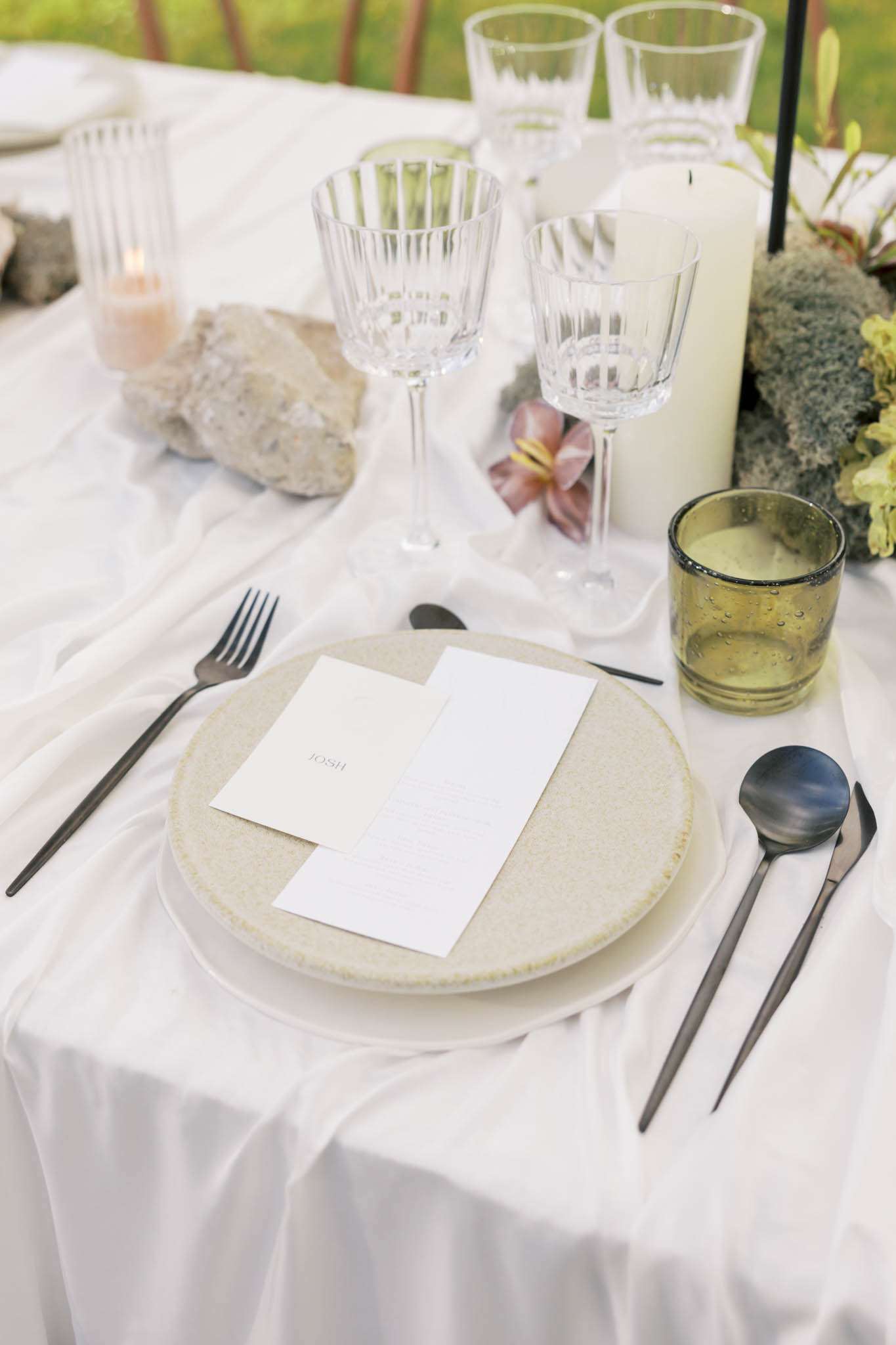 Close-up place setting with speckled stoneware plate, black cutlery, pillar candle, and pink lily on white linen