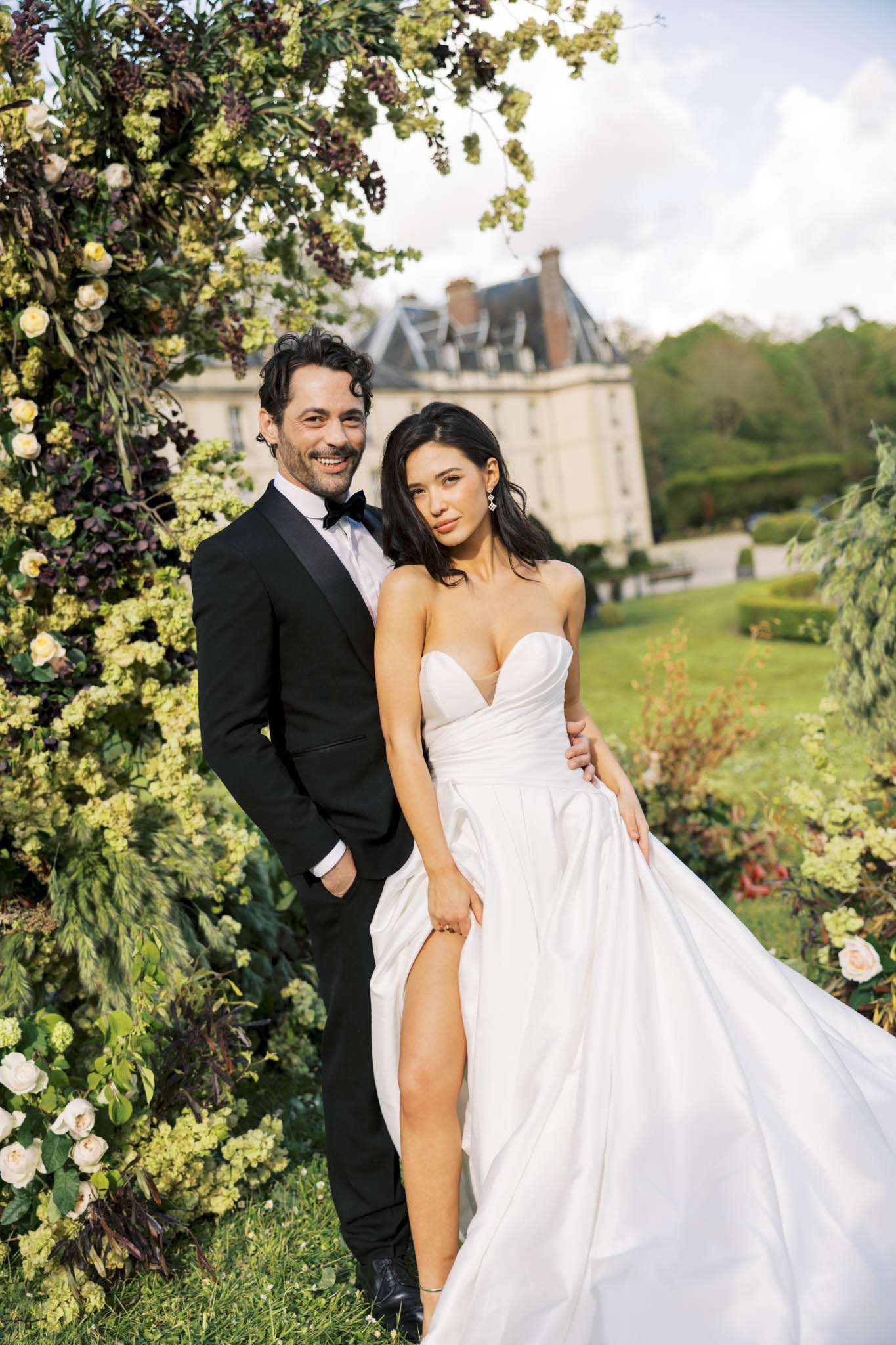 A couple portrait taken outdoors in the manicured grounds of a French château, which is visible in the background with its classic mansard roof and stone facade. The groom wears a black tuxedo with a bow tie, while the bride wears a white strapless sweetheart-neckline ballgown with a high front slit and a full draped skirt; she accessorizes with diamond drop earrings and holds the skirt of her dress to one side. They are posed in front of a large floral arch constructed with cream garden roses, chartreuse viburnum, and deep burgundy-black hellebores and foliage, creating a lush, dark-romantic botanical frame. The shot is a mid-length portrait with a shallow depth of field that keeps the château softly blurred in the background, with formal garden hedges visible on either side of the couple.