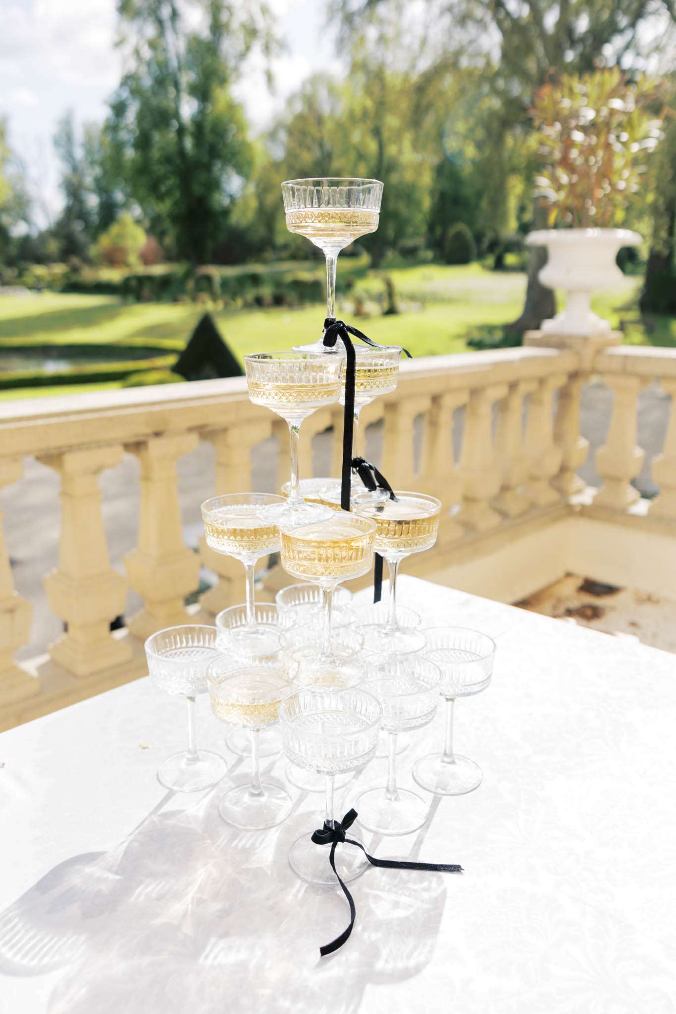 A champagne tower made up of cut crystal coupe glasses arranged in four tiers is displayed on a white linen-covered table during what appears to be a cocktail hour or reception. The glasses are partially filled with golden champagne and are tied together with black velvet ribbon bows at each tier, adding a classic black-and-white accent to the styling. The setup is positioned on an outdoor stone balustrade terrace, with a formal French-style garden visible in the background featuring manicured hedges and a decorative white stone urn. The shot is a close-up detail image taken at a slight angle, with the garden softly out of focus behind the tower.