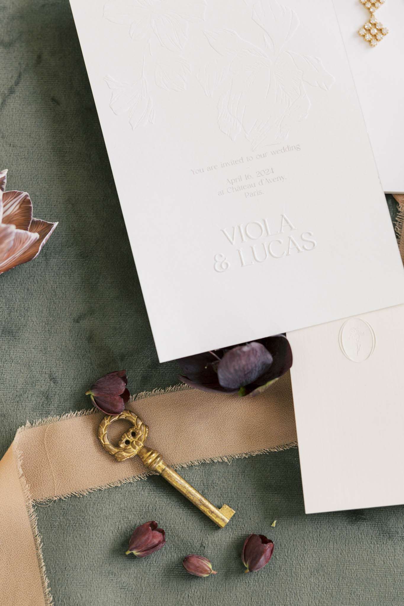 Blind-embossed ivory invitation with champagne ribbon, brass key, and burgundy tulip buds on sage velvet