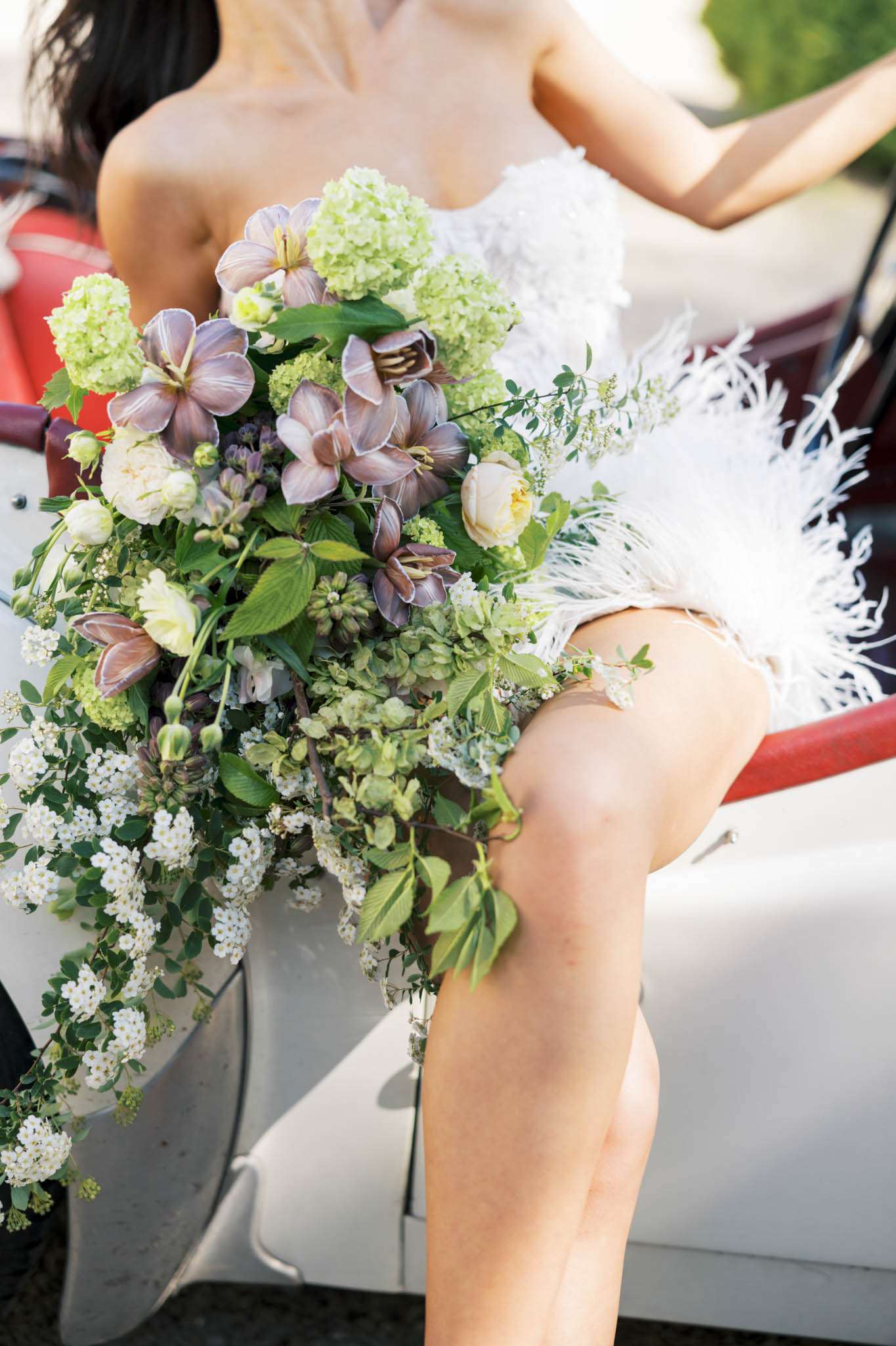 Bride in white feather-trimmed mini dress holding a cascading bouquet of purple clematis and cream roses in a vintage car
