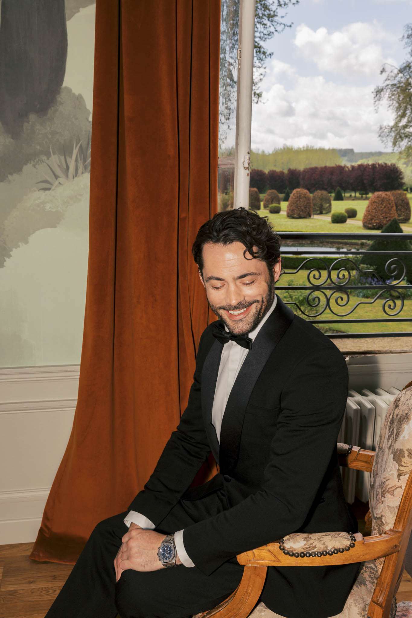 Groom in black tuxedo and bow tie seated in vintage armchair smiling, with chateau garden visible through window