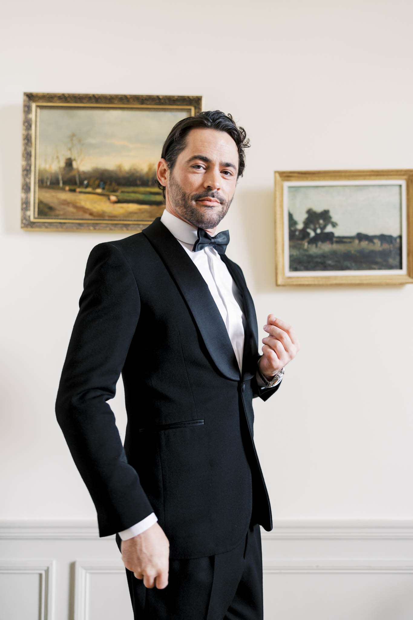 Groom in black tuxedo with satin lapels adjusting jacket in room with white wainscoting and oil paintings