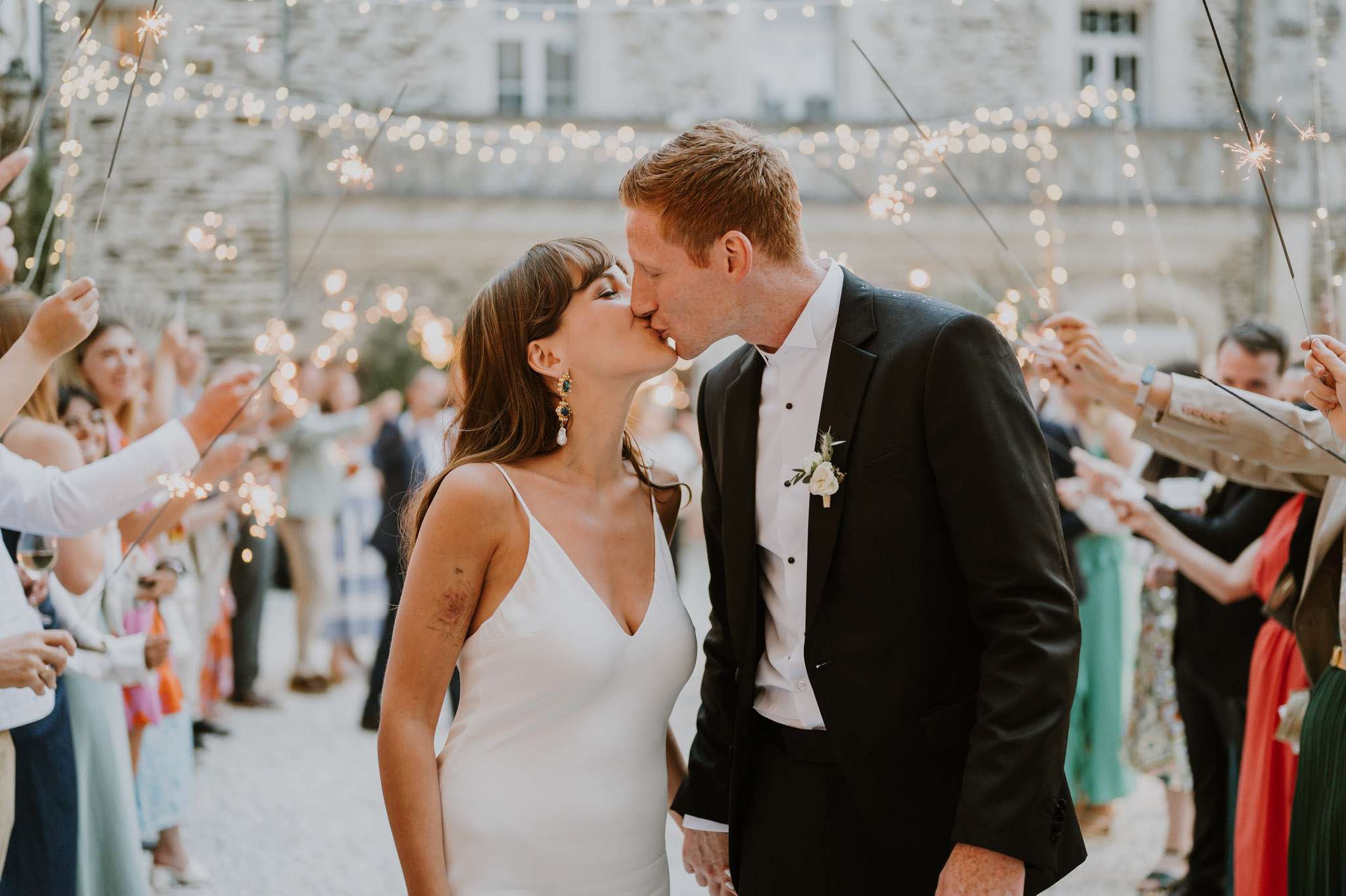 Couple kissing during sparkler send-off in chateau courtyard with thirty guests fairy lights and evening glow