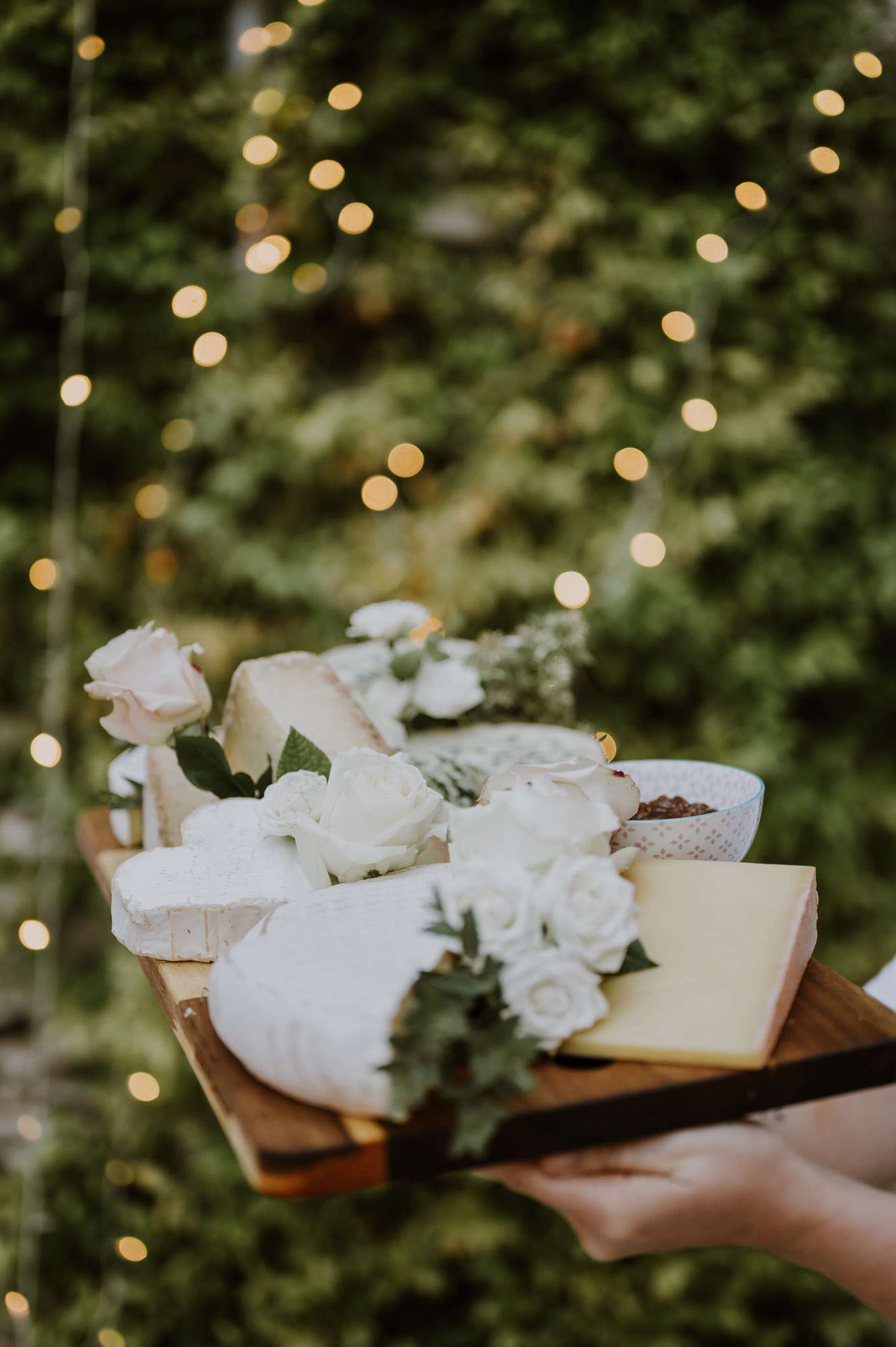 Wooden cheese board with French cheeses, white roses, and chutney held outdoors with fairy light bokeh