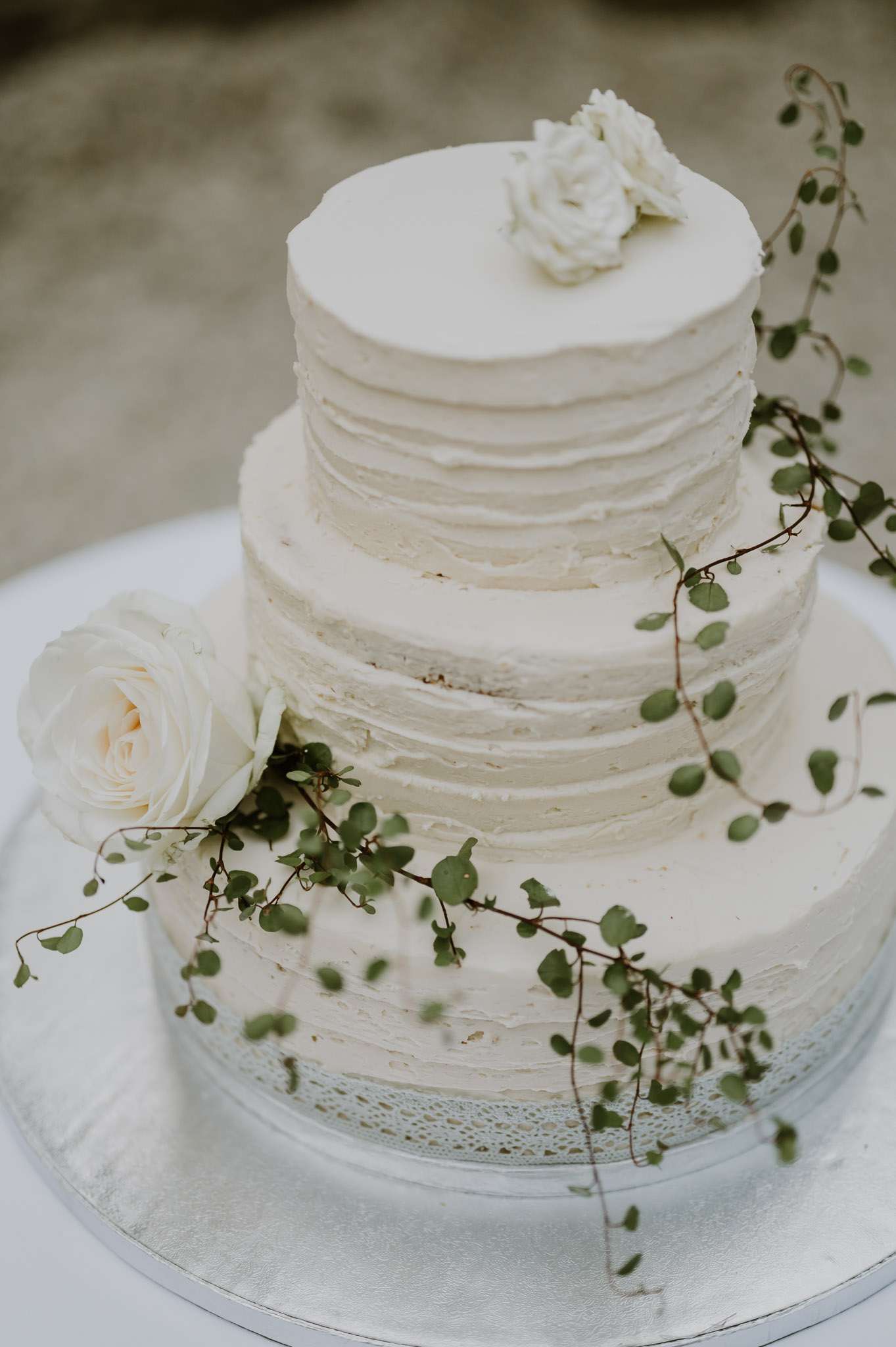 Close-up detail shot of a three-tiered semi-naked wedding cake with horizontally textured ivory buttercream frosting, placed on a silver cake stand with a lace doily on a white linen-covered table. The cake is decorated with trailing green vine sprigs with small rounded leaves cascading diagonally across all three tiers, one cream garden rose on the lower left tier, and a small white ranunculus or peony bloom on the top tier. The styling is organic and minimal, consistent with a natural or botanical aesthetic. The background is softly blurred, suggesting an outdoor setting.