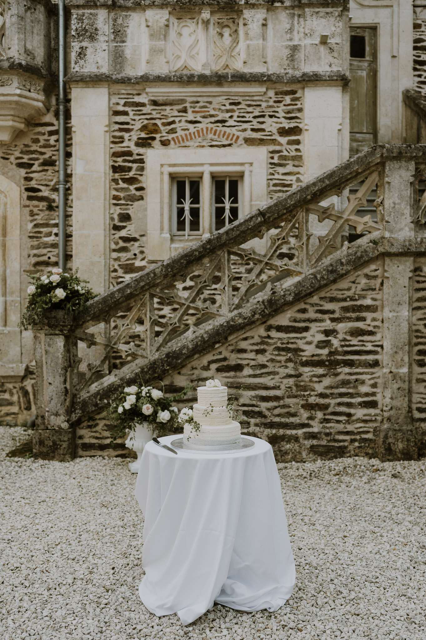 Three-tier white cake with trailing greenery and blush roses before Gothic stone chateau staircase