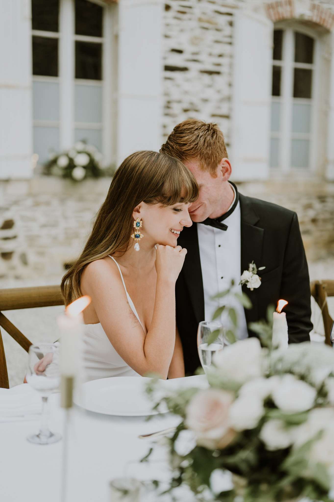 The bride and groom are seated together at what appears to be an outdoor reception dinner table set against the facade of a French château with white shuttered windows. The bride wears a white spaghetti-strap slip-style gown and gold statement earrings featuring blue gemstones and a pearl drop, while the groom is dressed in a black tuxedo with a bow tie and a white floral boutonniere. The table is dressed with a white linen, white ceramic plates, crystal glassware, and lit taper candles, with a soft blush and white floral centerpiece with greenery visible in the foreground. The overall styling is classic and understated, and the shot is a close-up portrait framing the couple from approximately the waist up.
