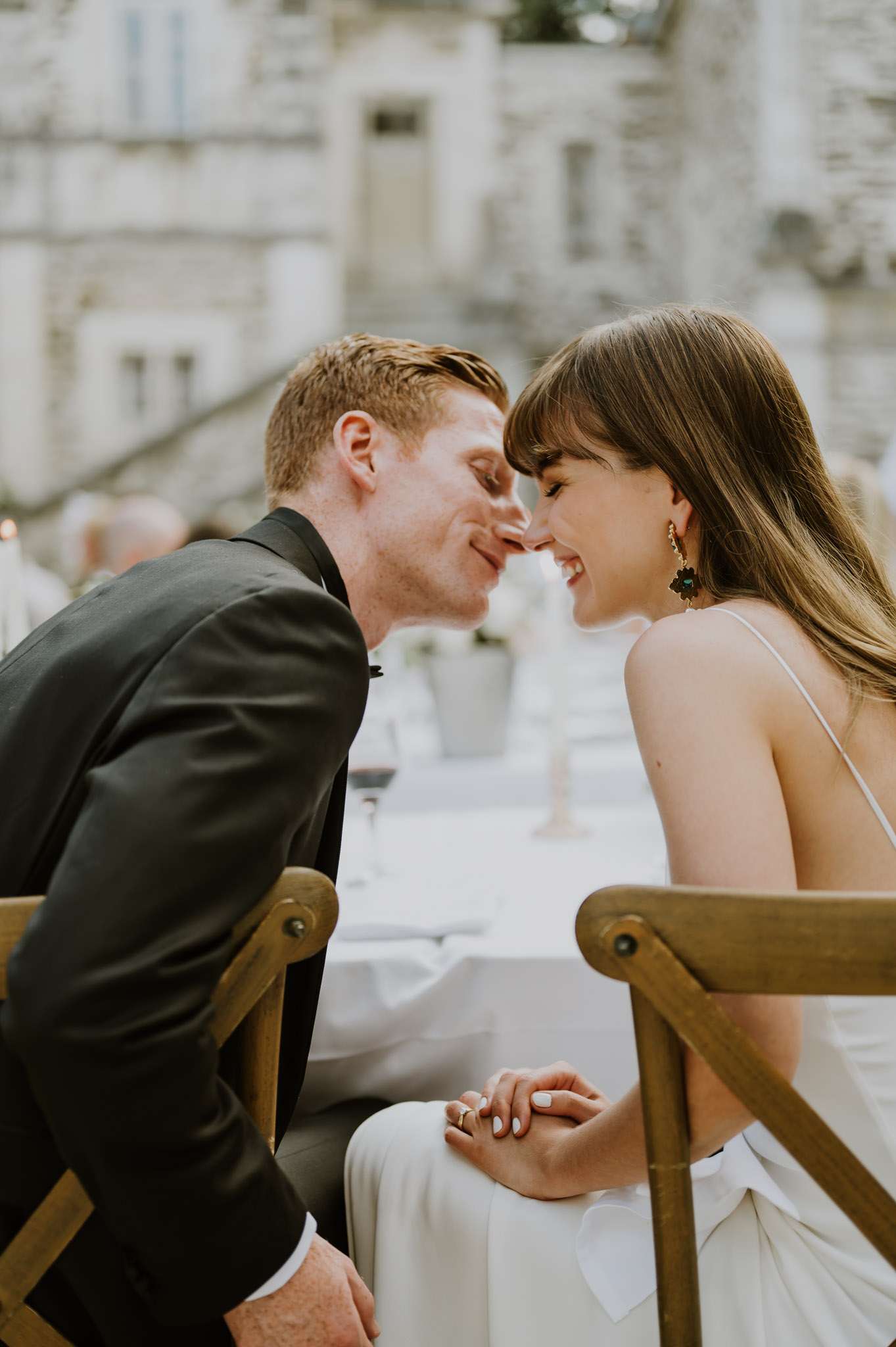 Couple touching foreheads at outdoor reception table, bride in minimal spaghetti-strap gown with dark earrings