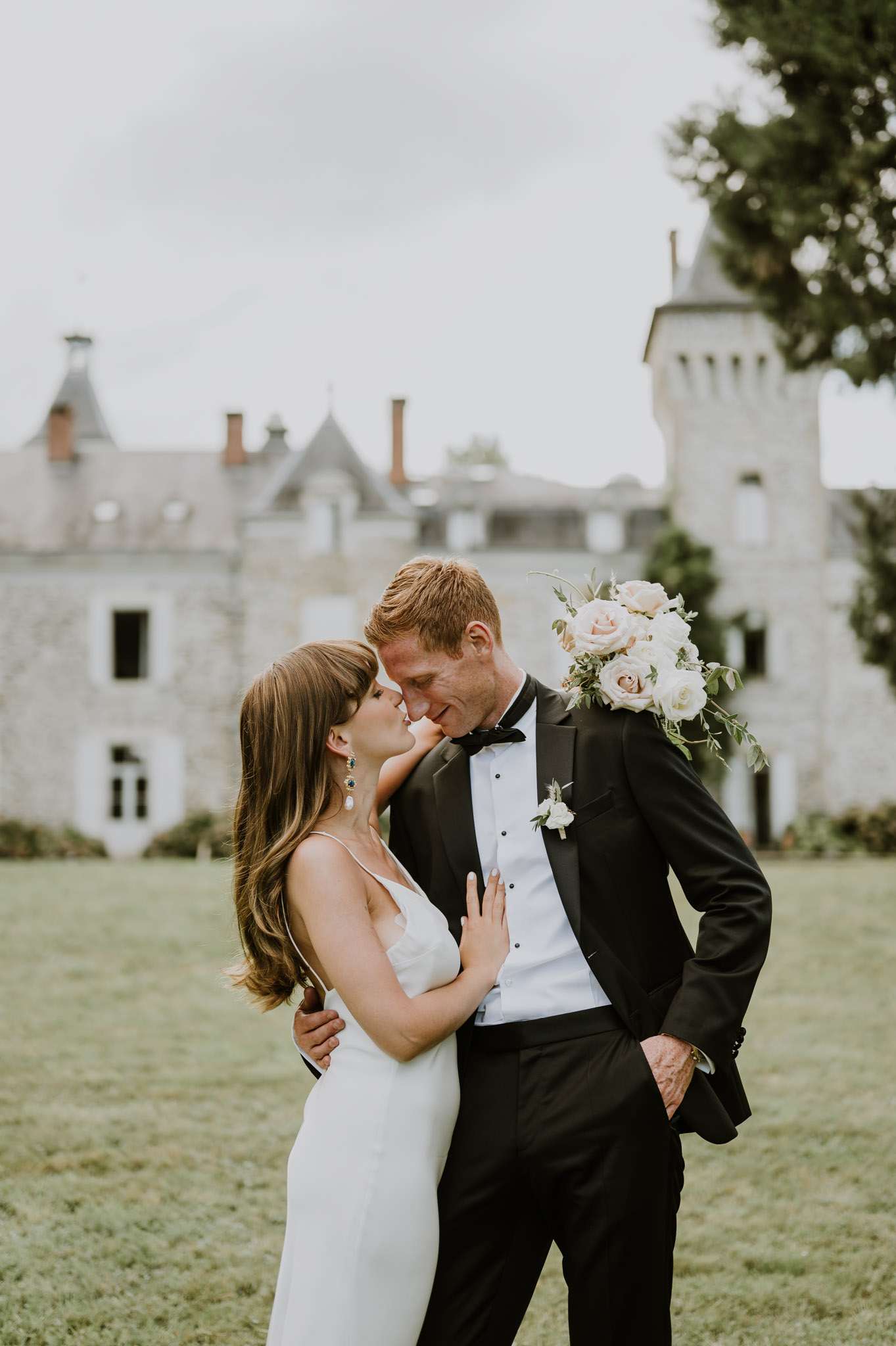 An outdoor couple portrait taken on the grounds of a French château, with the stone castle building and tower visible and softly out of focus in the background. The bride wears a fitted ivory slip-style gown with thin spaghetti straps and a deep V-neckline, paired with drop earrings featuring dark gemstones; she holds a bouquet of blush and white garden roses with trailing greenery. The groom wears a classic black tuxedo with a bow tie and a small white boutonnière, with one hand in his pocket. The two are posed close together, foreheads nearly touching in an intimate moment, shot as a mid-length portrait with a slightly warm, muted color tone.