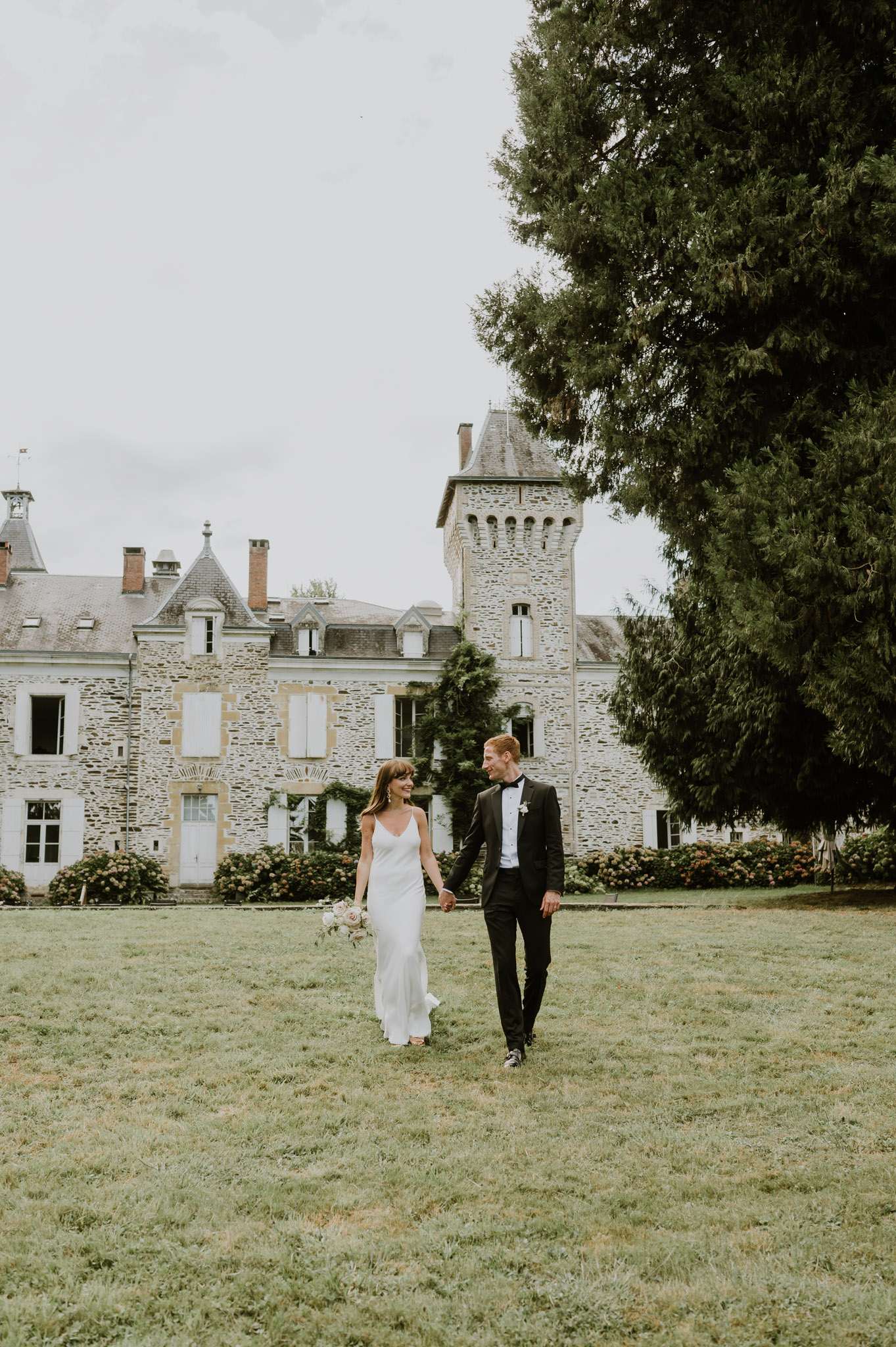 Couple walking hand in hand on chateau lawn bride in ivory slip gown with blush bouquet groom in tuxedo tower behind