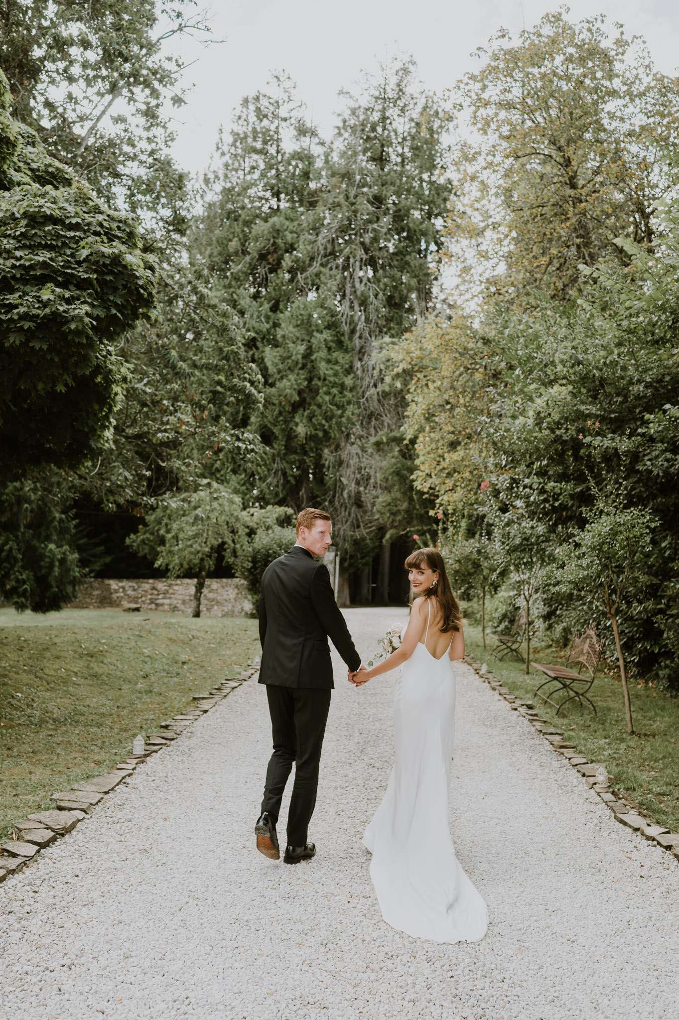 A couple portrait taken outdoors on a gravel path lined with stone edging, set within the grounds of what appears to be a French estate or château garden. The groom wears a fitted black suit with black dress shoes and walks slightly ahead, while the bride turns back toward the camera with a small smile, holding his hand. She wears a minimalist ivory slip-style gown with thin spaghetti straps and a low open back, with a short train trailing on the gravel, and carries a small white bouquet. The overall styling is modern and minimal. The shot is a wide, full-length portrait taken from a slightly elevated angle along the central axis of the path.