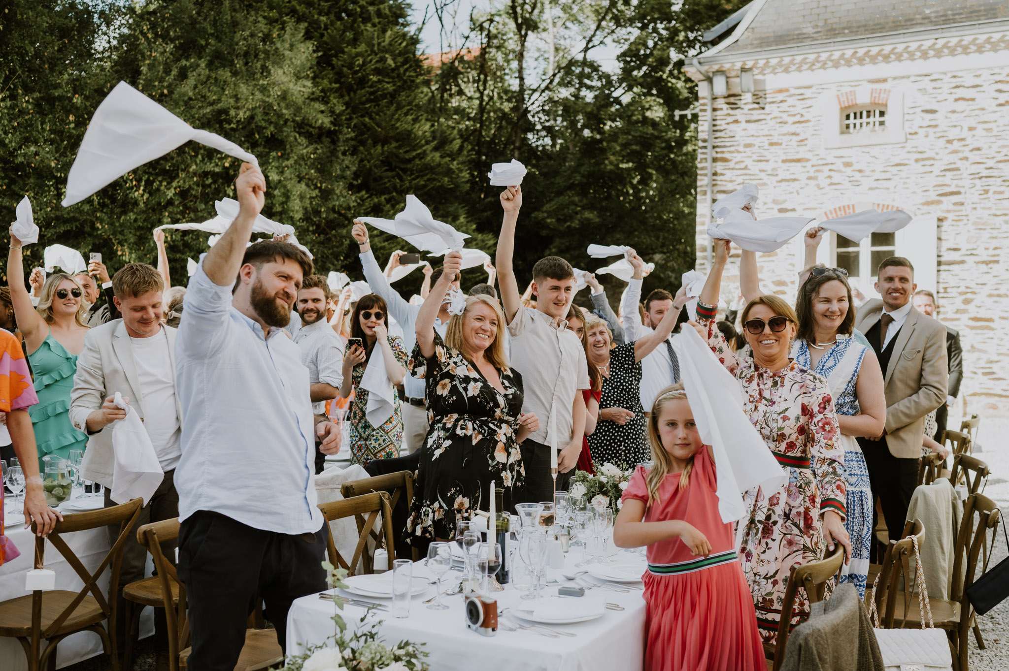 Wedding guests waving white napkins at outdoor reception dinner beside stone French country house