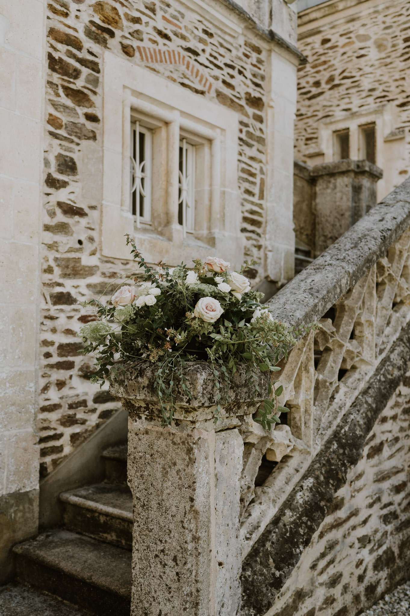 A close-up detail shot of a floral arrangement placed on top of a stone newel post at the base of an outdoor staircase at what appears to be a French château. The arrangement features blush and ivory roses, white lisianthus or similar small white blooms, queen anne's lace, and abundant loose greenery including eucalyptus and trailing foliage, styled in an unstructured, garden-gathered aesthetic. The stone balustrade with decorative cutwork detailing and the mixed rubble and ashlar stone façade of the building are visible in the background. No people are present. Potential venue feature image.