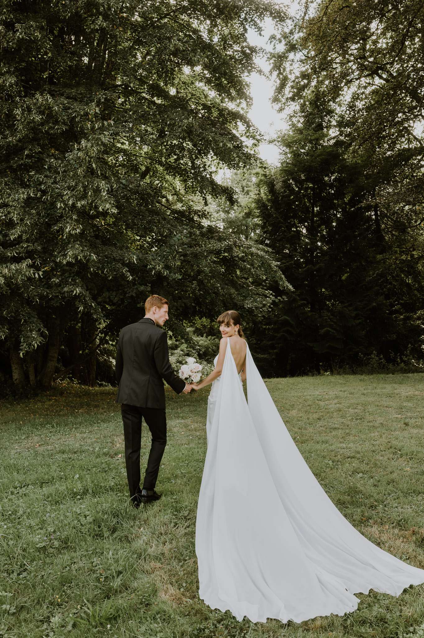 Bride and groom walking hand-in-hand across a lawn with bride's cape train flowing behind them