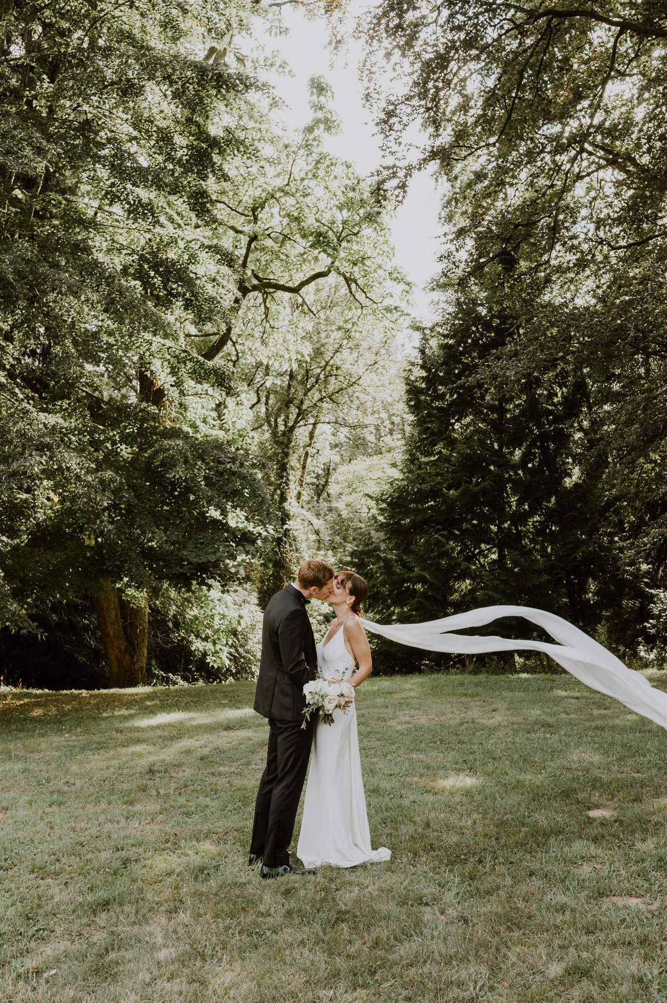 A couple portrait taken outdoors on a lawn surrounded by tall mature trees, with the bride and groom kissing. The groom wears a dark navy or black suit, and the bride wears a sleek, minimalist ivory slip-style gown with a halter neckline and a long white veil that is caught by the wind and billows dramatically upward and to the right of the frame. The bride holds a loose bouquet of blush and white blooms with greenery. The overall styling is modern and minimal. The shot is a full-length portrait with the tree canopy framing the couple from above.