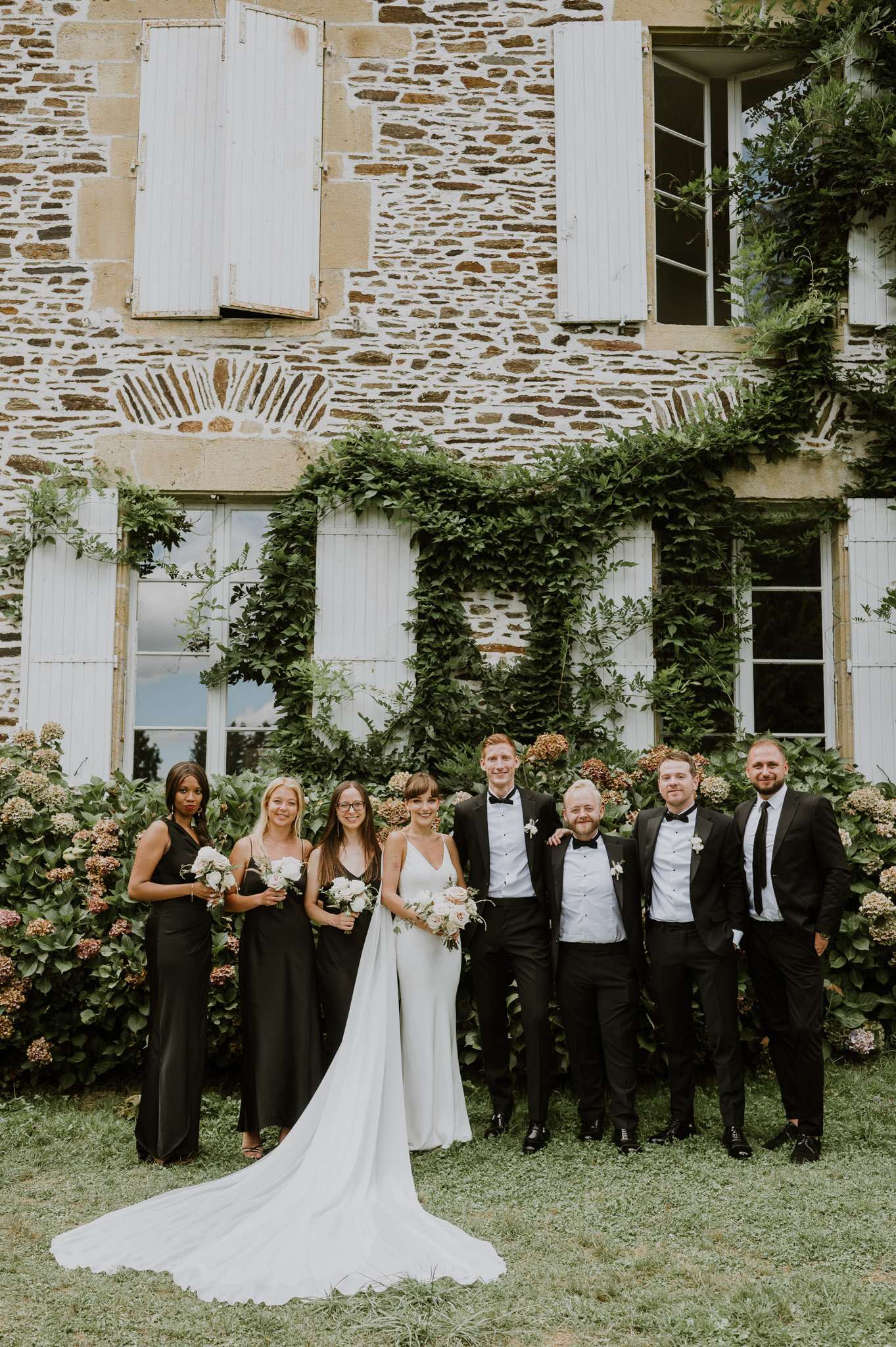 A bridal party portrait taken outdoors in front of a two-storey French stone manor house with white shutters and climbing green foliage. The group of seven stands on a lawn in front of hydrangea bushes with muted blush and dried-flower tones. The bride wears a simple ivory slip-style gown with a long train and holds a bouquet of white and blush blooms; the three bridesmaids wear floor-length black dresses and carry small white floral bouquets. The four groomsmen and groom are dressed in black tuxedos with bow ties, with one man wearing a dark suit and tie instead. The shot is a full-length wide portrait with the full group posed in a single row facing the camera.