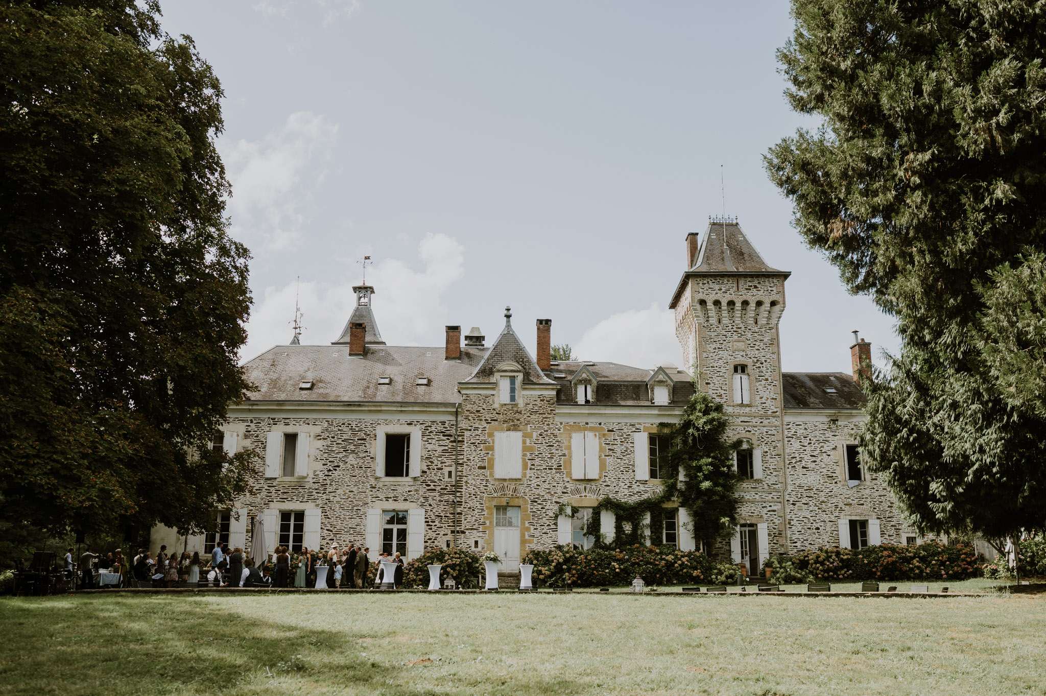 A wide shot of a French château during what appears to be an outdoor cocktail hour, with a crowd of approximately 50–80 guests gathered along the front façade of the building. The château is a multi-story stone structure with slate-roofed conical towers, white shuttered windows, climbing vines, and flowering hydrangea bushes along the base. White cocktail tables and tall poseur tables are visible among the guests. The foreground shows a large open lawn. Potential venue feature image.
