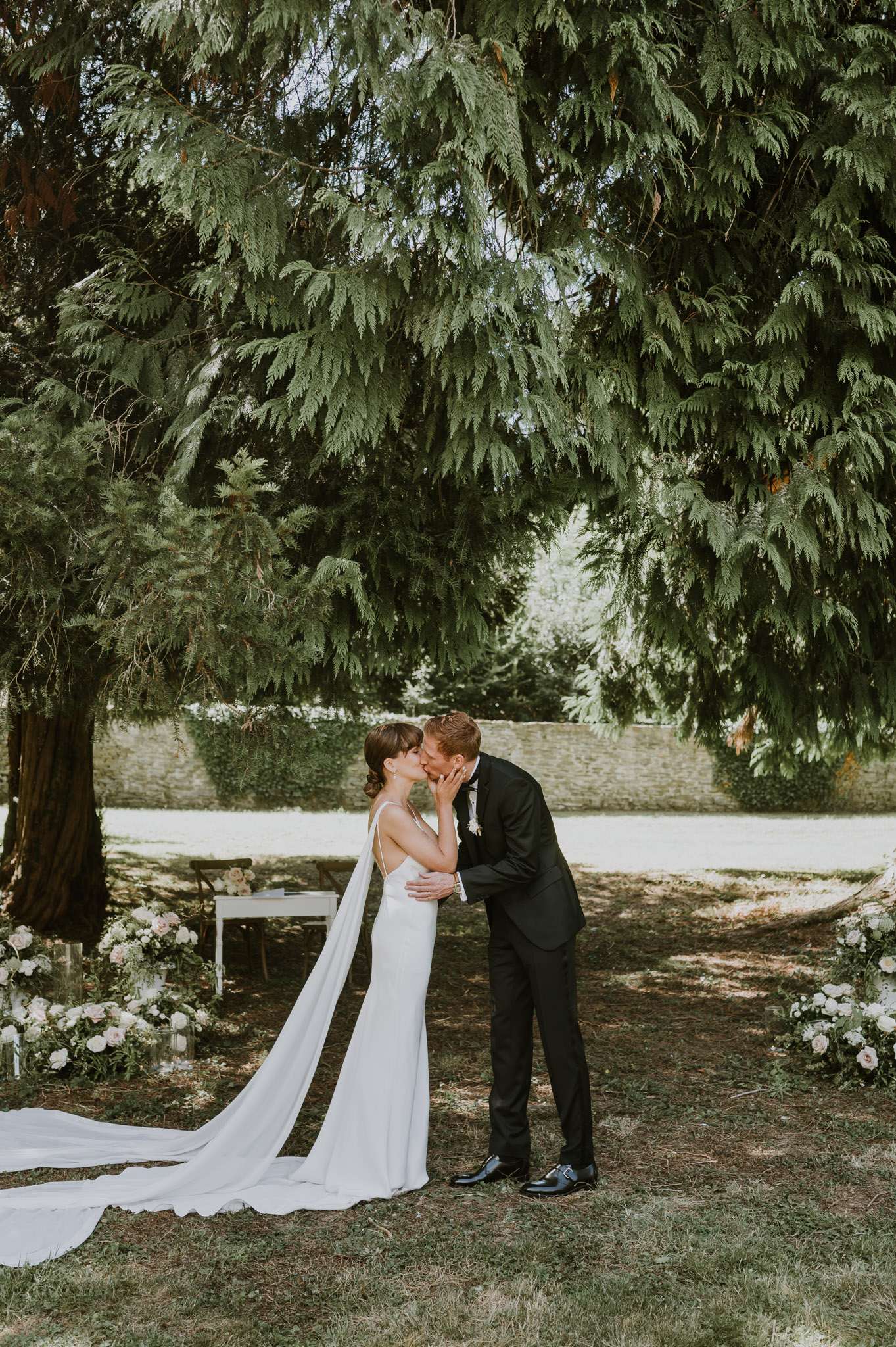 The bride and groom share their first kiss during an outdoor ceremony held beneath a large tree in a walled garden setting. The bride wears a fitted ivory slip-style gown with thin spaghetti straps, a low open back, and a long cathedral-length veil that trails across the ground; the groom wears a black tuxedo with a bow tie and a white floral buttonhole. A white signing table with dark wood chairs is visible in the background, flanked by low ground-level floral arrangements featuring blush and white roses with lush greenery, placed on either side of the ceremony space. The composition is a full-length portrait shot, with the couple centered in the frame.