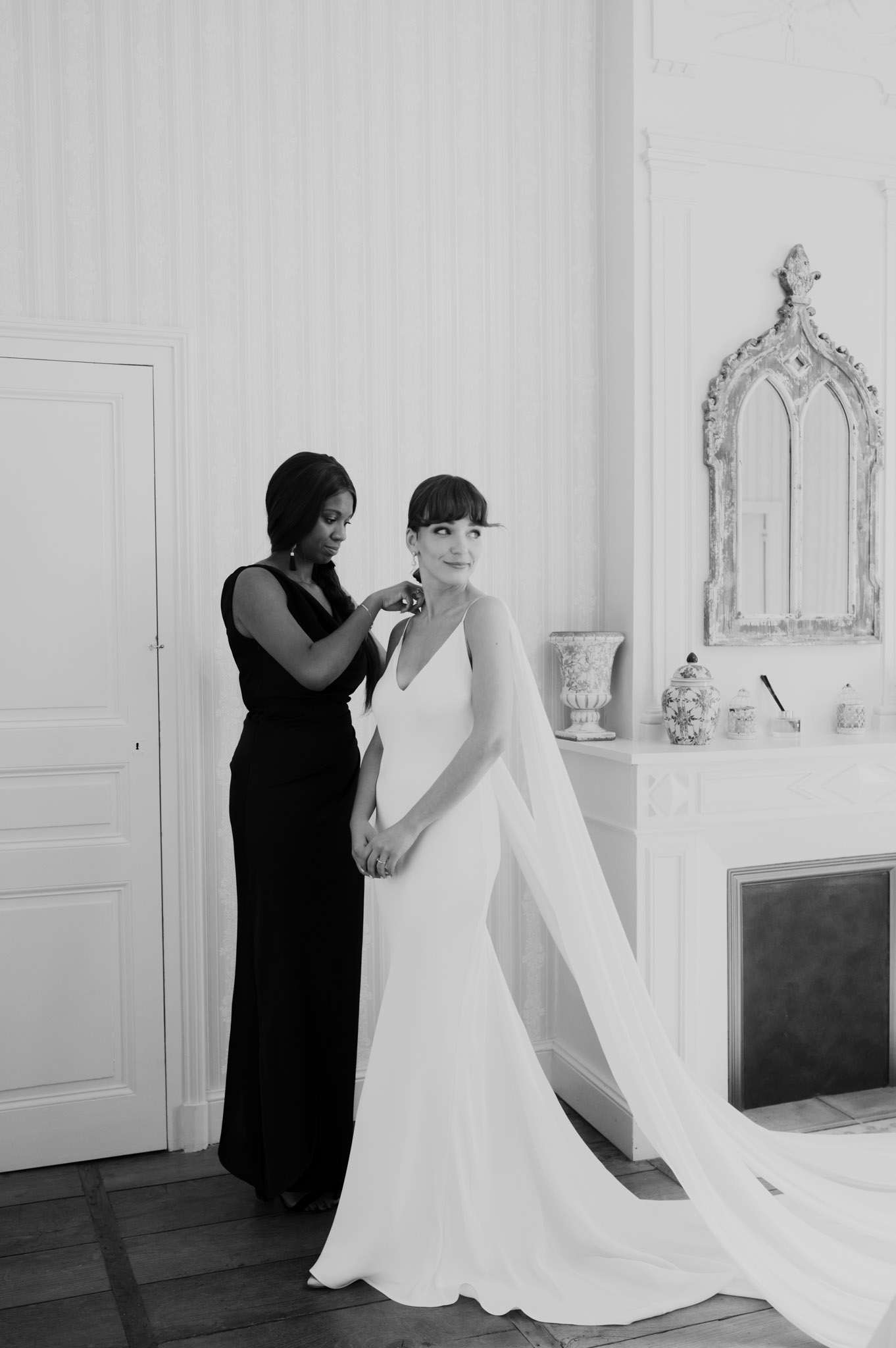 Black and white shot of woman helping bride fasten earring in chateau room with ornate mirror and fireplace
