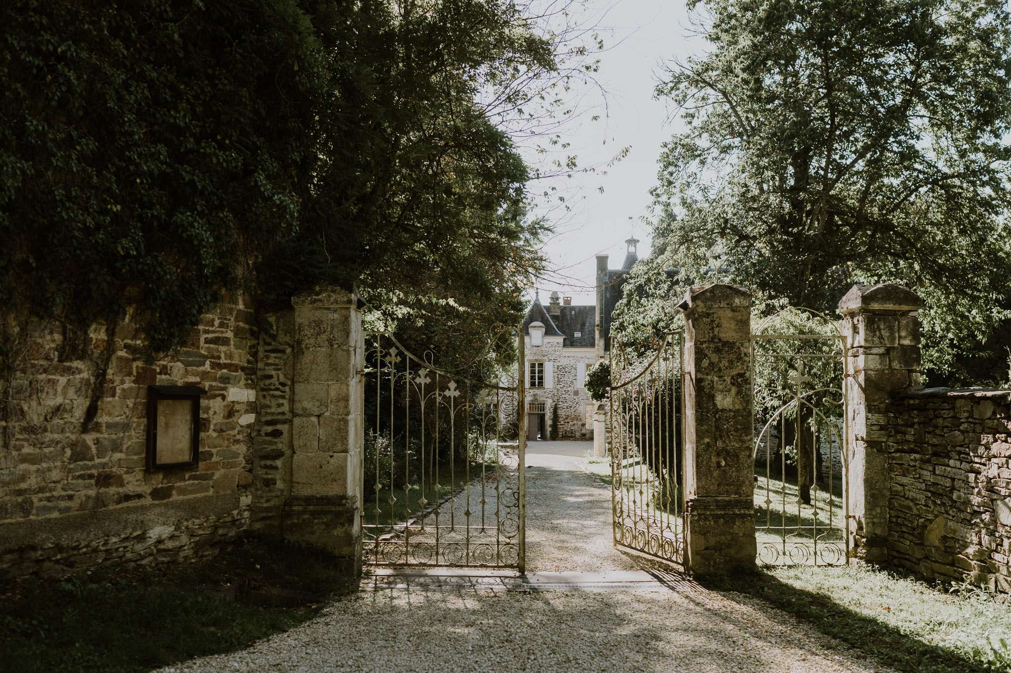 A wide shot looking through a set of open ornate wrought-iron gates mounted between aged stone pillars, leading down a gravel driveway toward a French manor or château in the background. The property features slate-roofed turrets and stone façade typical of historic French country architecture. No people are visible in the frame. The image has a slightly muted, warm tonal quality with dappled natural light filtering through the surrounding trees. Potential venue feature image.
