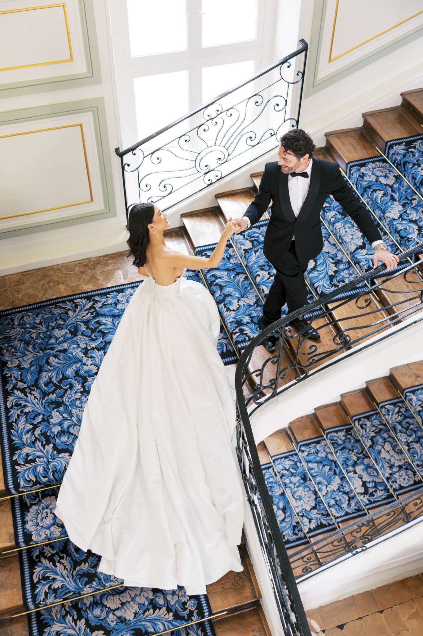 Overhead portrait of bride and groom holding hands on ornate chateau staircase with blue damask carpet runner