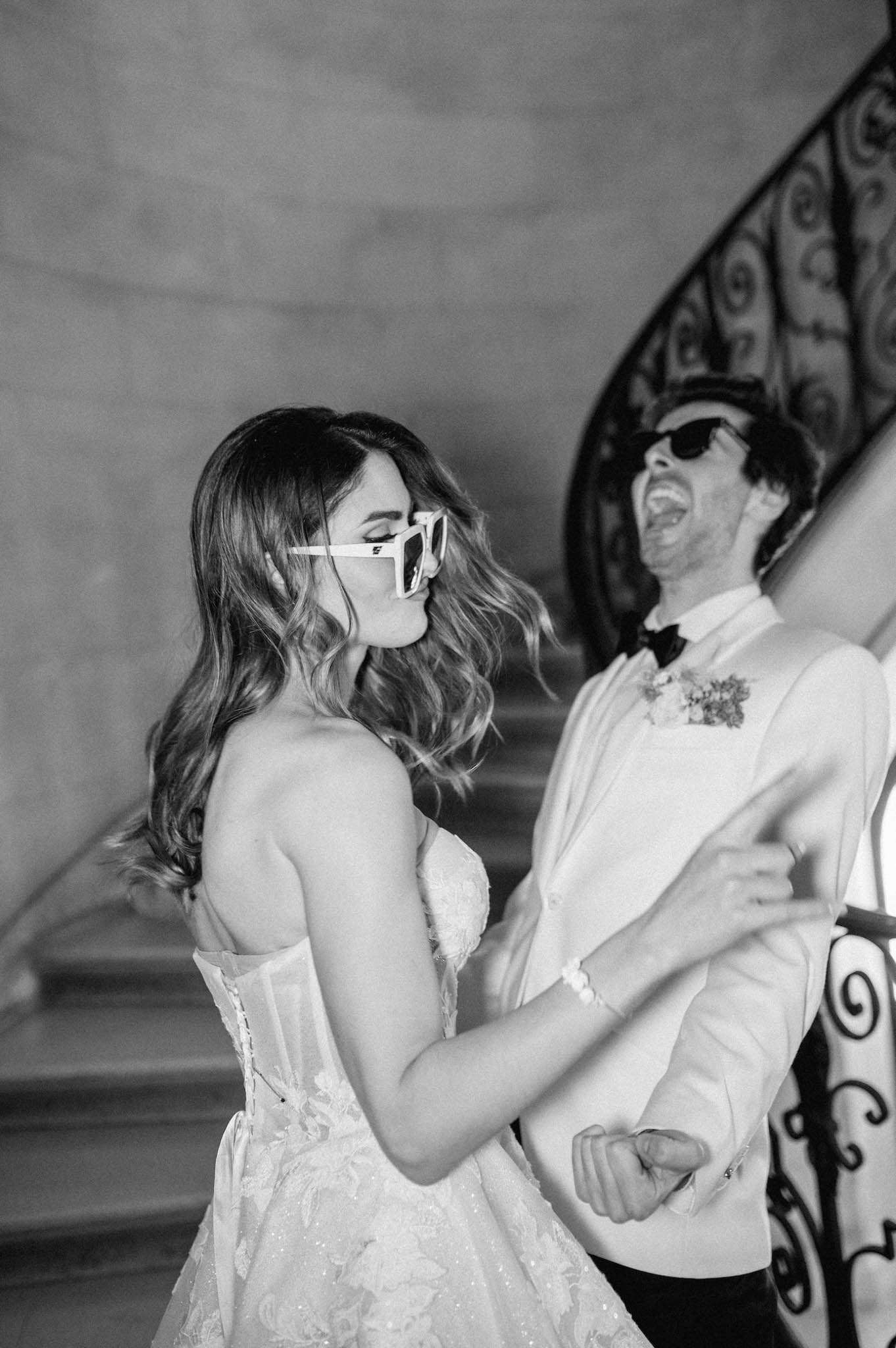 A black-and-white portrait of a bride and groom laughing and dancing together on an indoor staircase landing, likely during a reception moment. The bride wears a strapless lace corset-bodice gown with wavy, loose hair and oversized white-framed sunglasses; the groom wears a white dinner jacket with a black bow tie, a floral boutonnière, and dark sunglasses. The setting features a curved wrought-iron staircase railing in the background, suggesting a château or grand manor interior. The image is shot at medium range with soft, even light and strong mid-tone contrast typical of film-style black-and-white photography.