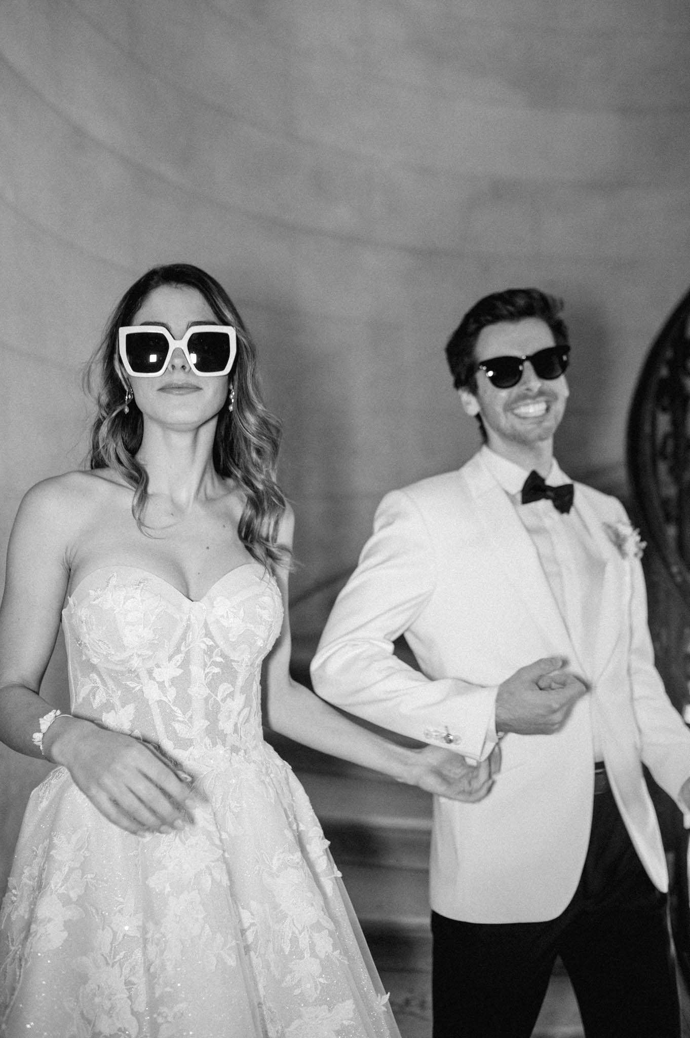 Black and white portrait of bride in lace ballgown and groom in white dinner jacket wearing sunglasses and laughing