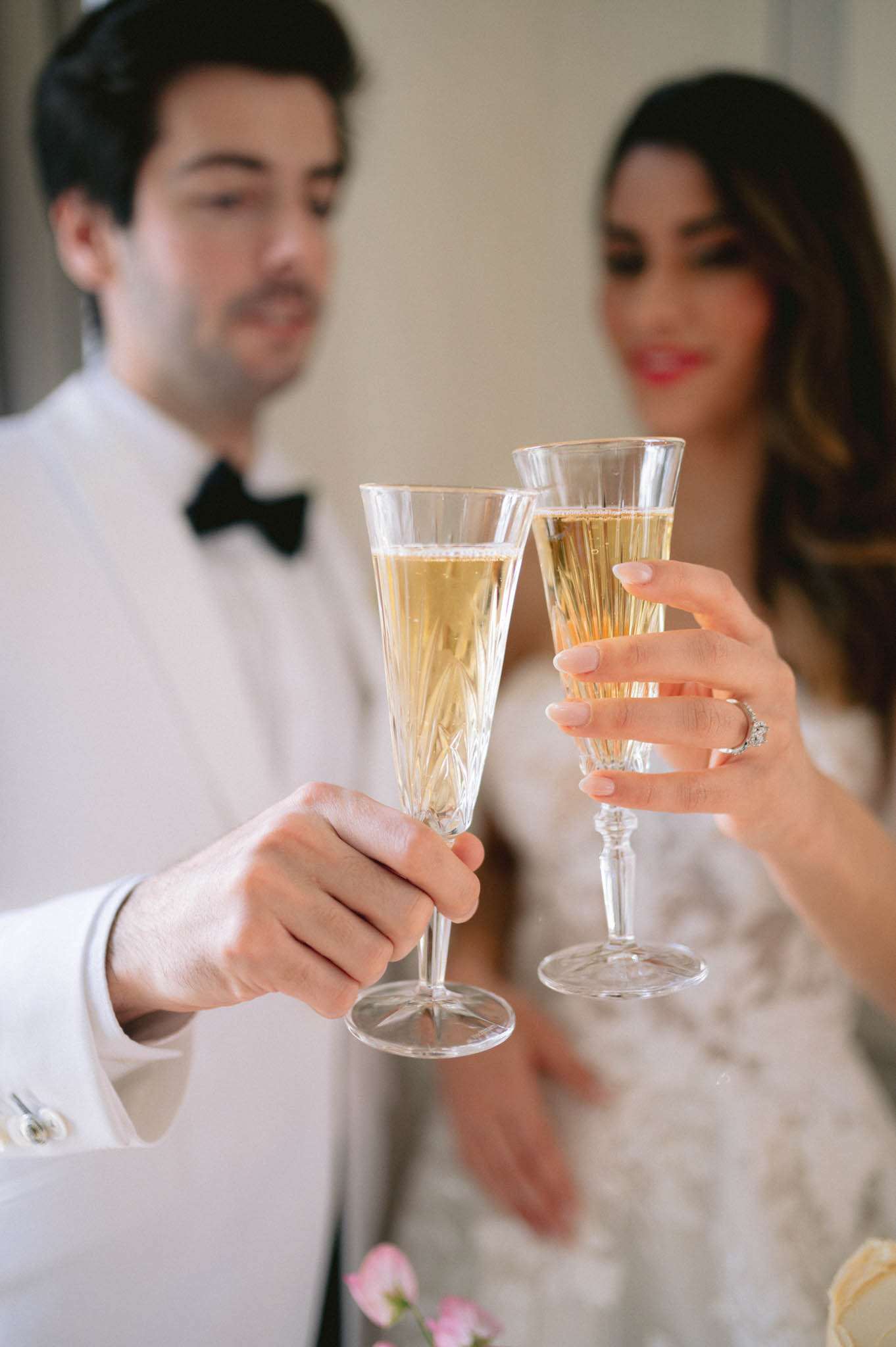 The bride and groom are toasting with crystal champagne flutes filled with golden sparkling wine, in what appears to be an indoor setting with a neutral background. The groom wears a white dinner jacket with a black bow tie, while the bride wears a white lace or embroidered gown and has a diamond engagement ring visible on her finger with pale pink manicured nails. The bride has long dark hair with highlights and is wearing red lipstick, smiling toward the groom. The composition is a close-up portrait with the champagne flutes sharp in the foreground and the couple softly blurred in the background, with a hint of pink and cream florals at the bottom edge of the frame.