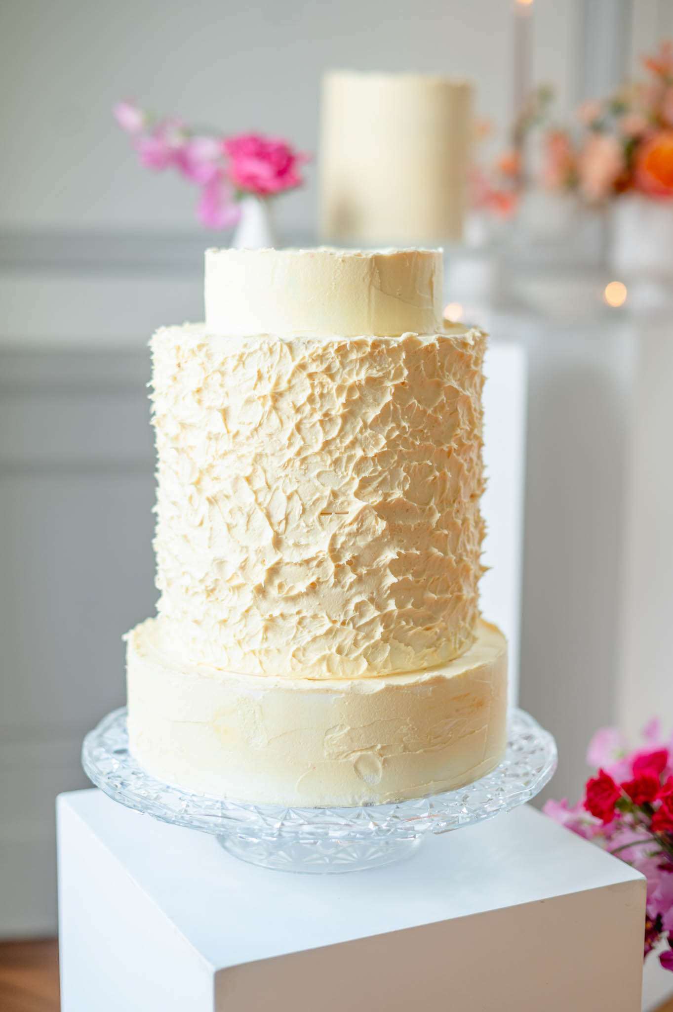 Three-tier ivory buttercream cake with textured palette-knife middle tier on crystal stand with hot pink florals behind