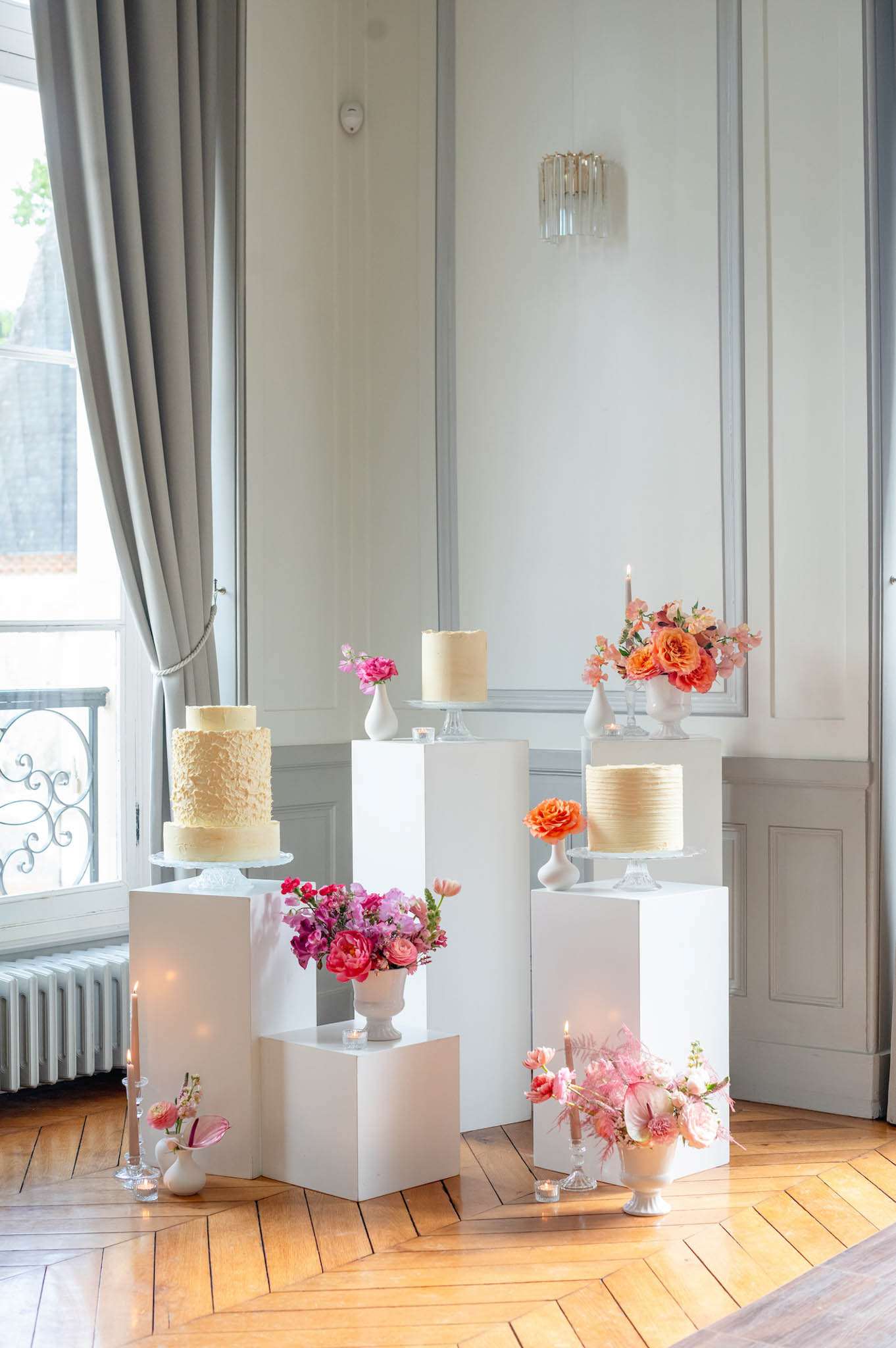 A styled cake display is arranged indoors in a room with white panelled walls, a crystal wall sconce, grey floor-length curtains, and herringbone parquet flooring. Five white-frosted wedding cakes of varying heights and textures — including a two-tier ruffled cake, a smooth ribbed single-tier, and a textured buttercream single-tier — are placed on white rectangular plinths and glass cake stands at different heights. The display is styled with multiple white urn and bud vase floral arrangements featuring hot pink ranunculus, magenta sweet peas, coral and orange garden roses, and blush pink blooms, distributed across the plinths and floor. Taper candles in glass holders and scattered tea lights are placed throughout the arrangement, adding warm accent lighting. The overall decor palette is white, coral, hot pink, and orange, with a clean modern-classic styling approach. Wide shot capturing the full installation.