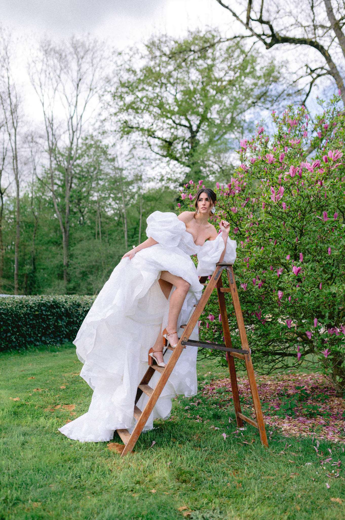 Bride in ruffled puff-sleeve ball gown posed on rustic wooden ladder beside magnolia tree
