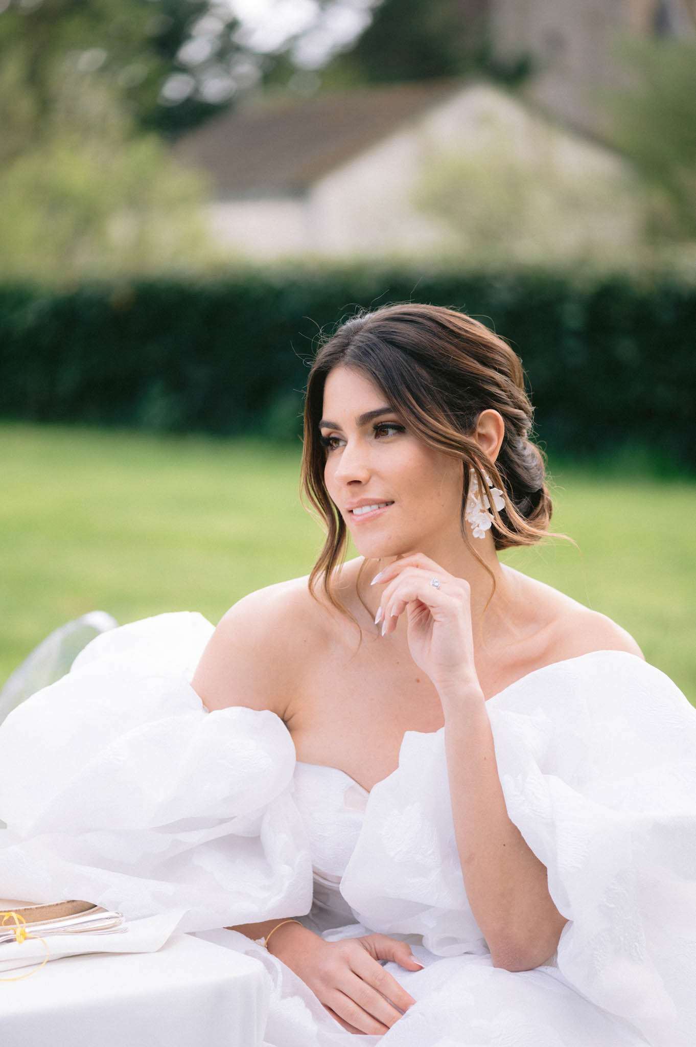 A close-up portrait of a bride seated at an outdoor table, set against a softly blurred garden and stone building background. She wears an off-the-shoulder white ballgown with voluminous puff sleeves and a sweetheart neckline, paired with floral drop earrings in cream and gold tones and a delicate gold bracelet. Her dark hair is styled in a low chignon with face-framing loose pieces, and her makeup features defined brows, subtle eye makeup, and a natural lip. She rests her chin lightly on her hand, gazing to one side, with a solitaire engagement ring visible on her finger.