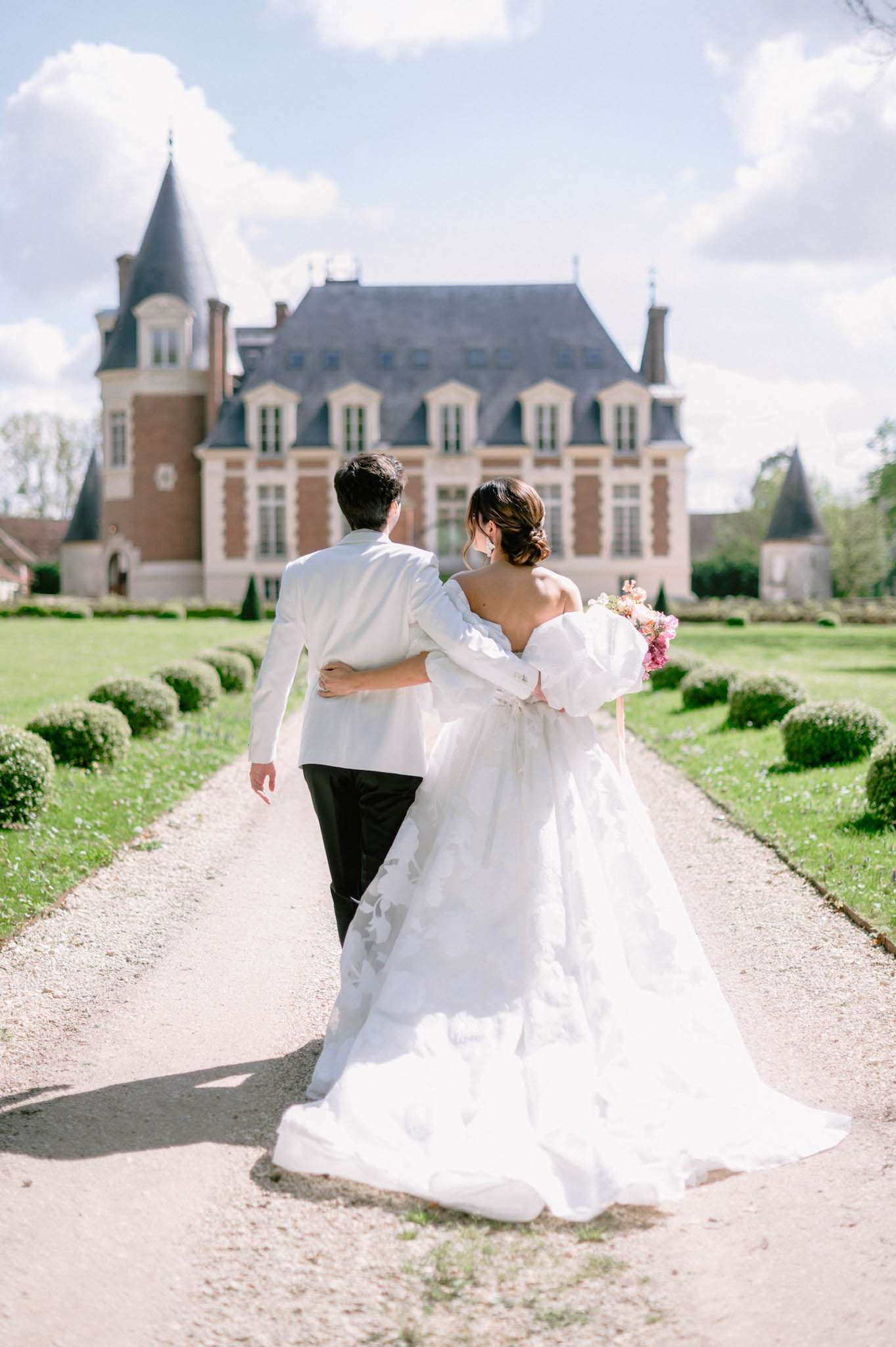 A couple portrait taken outdoors at a French château, showing the bride and groom walking away from the camera along a gravel path toward the main building. The groom wears a white dinner jacket with black trousers, and the bride wears a voluminous off-the-shoulder white ballgown with floral appliqué detailing and a long train; her hair is styled in a low updo. She carries a lush bouquet of pink and coral flowers. The formal garden flanking the path features neatly clipped round box hedges arranged symmetrically. The château in the background is a classic French manor house with brick and stone facade, steep slate mansard roofs, and conical corner turrets. Wide shot, taken from behind the couple with the venue centered in the background. Potential venue feature image.