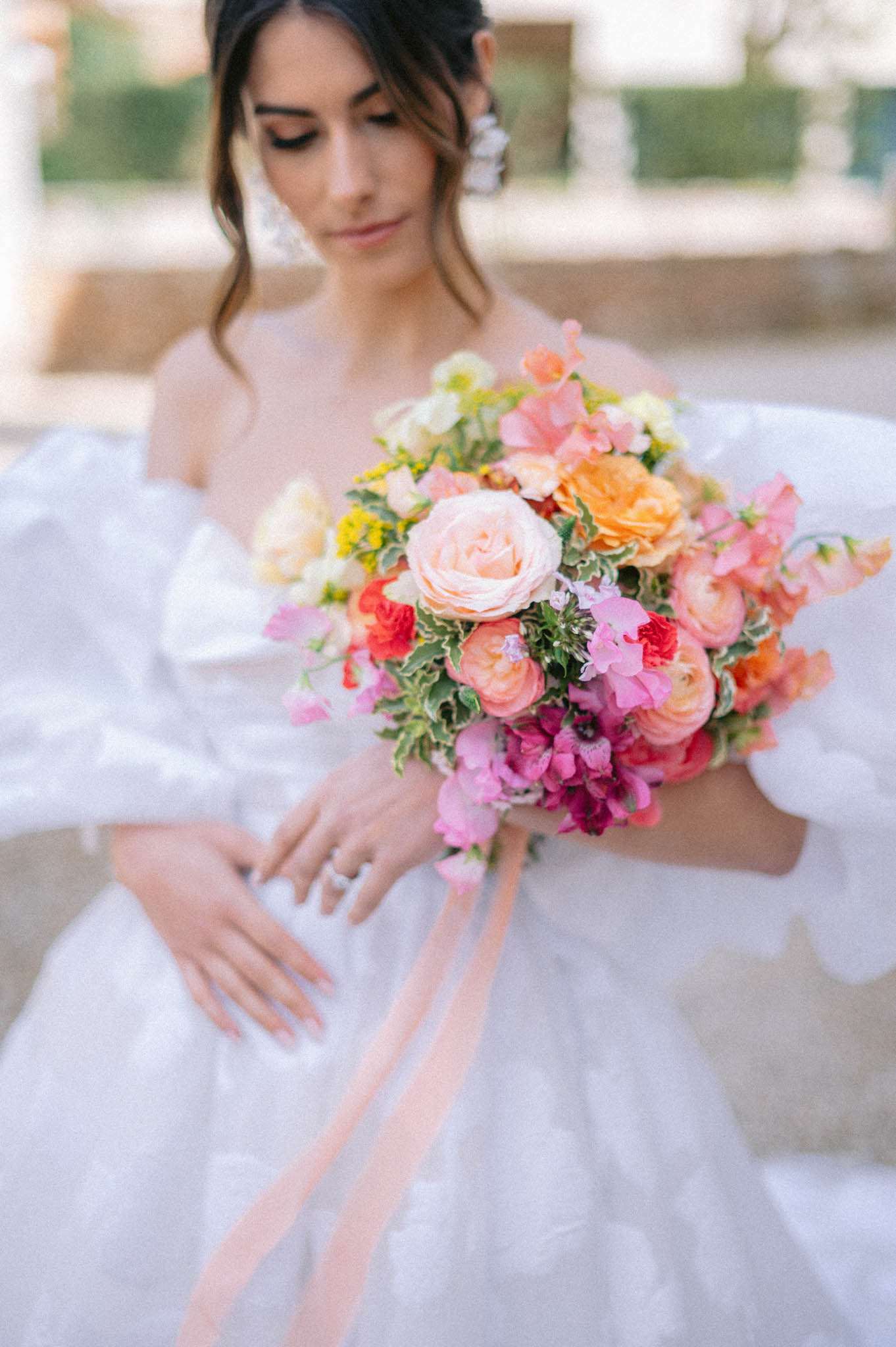 A bridal portrait shot outdoors, focused on a bride holding a vibrant, colorful bouquet while looking downward. She wears a white ball gown with voluminous puff sleeves and large floral drop earrings, with her dark hair pulled back in a loose updo. The bouquet is a dense, garden-style arrangement featuring peach garden roses, orange ranunculus, coral and hot pink sweet peas, magenta anemones, red accent blooms, yellow filler flowers, and variegated greenery, finished with long trailing blush silk ribbons. A solitaire diamond ring is visible on her hand. The composition is a close-up portrait with the bouquet as the primary focal point and the bride's face softly out of focus in the background.