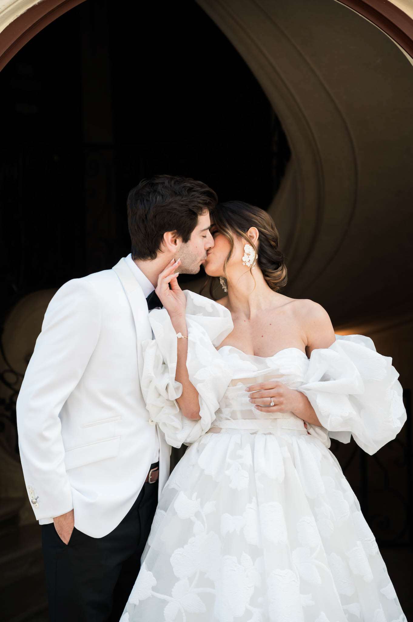A couple portrait showing the bride and groom kissing in front of a large dark archway or curved architectural structure that frames them from behind. The groom wears a white dinner jacket with black trousers and a black bow tie, while the bride wears a white strapless ball gown with a floral jacquard or appliqué skirt and voluminous detachable off-the-shoulder ruffle sleeves in organza. The bride has her dark hair styled in an updo and wears large white floral statement earrings; a diamond engagement ring and wedding band are visible on her hand. The composition is a mid-length portrait shot with the dark circular arch creating strong contrast against the white of both outfits, giving a classic, high-contrast look. The overall styling reads as modern formal with bold, sculptural bridal details.