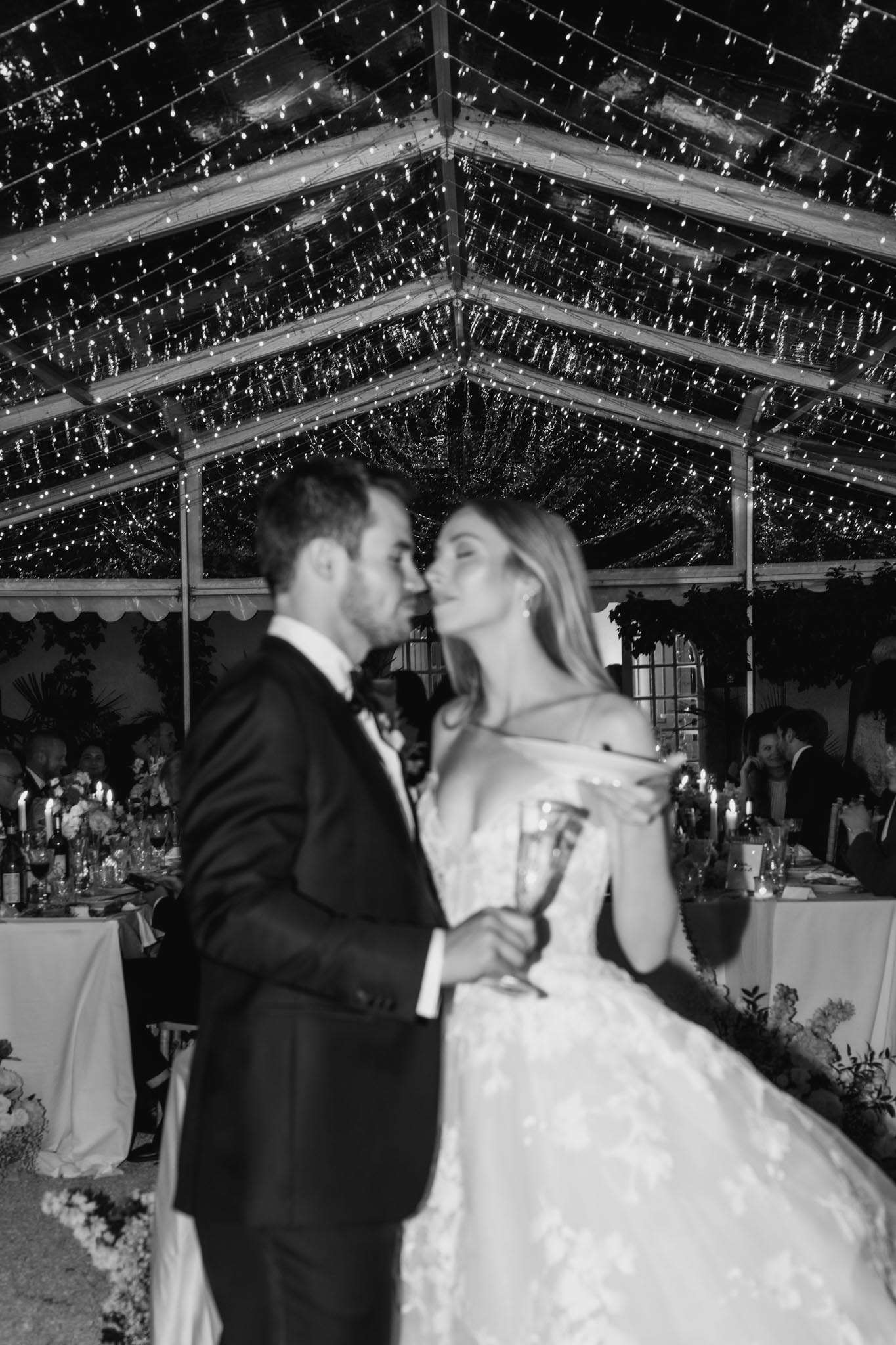 This black-and-white image shows a bride and groom sharing an intimate moment during their wedding reception, likely just after or during the first dance. The couple is slightly out of focus in the foreground — the groom wears a dark tuxedo with a bow tie, and the bride wears an off-the-shoulder ball gown with floral appliqué detailing; she holds a champagne flute. The setting is a clear-roofed marquee or greenhouse-style tent densely covered with fairy lights across the entire ceiling structure, creating bright, high-contrast sparkle in the upper half of the frame. In the background, reception dining tables are visible set with candles, floral centerpieces, and glassware, with seated guests visible on both sides. The composition is a mid-to-close portrait shot with shallow depth of field, drawing focus to the fairy-light canopy overhead while the couple remains softly blurred in the foreground.