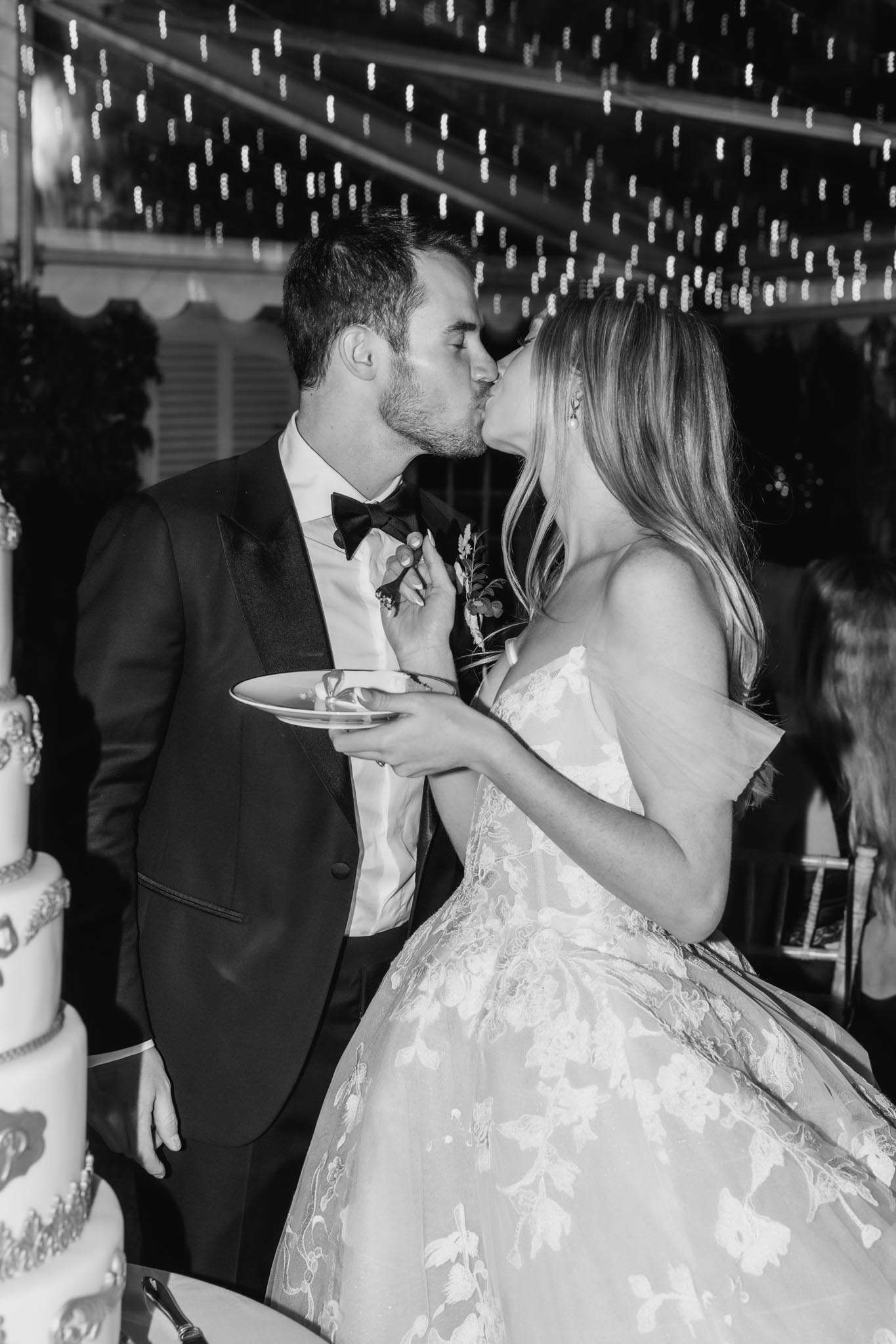 This black-and-white image captures the couple kissing immediately after the cake cutting during an indoor evening reception. The groom wears a dark tuxedo with satin lapels and a black bow tie, with a small floral boutonniere featuring greenery; the bride wears an off-the-shoulder gown with an all-over floral lace appliqué pattern and a full ballgown skirt. The bride holds a small plate with a slice of wedding cake while the multi-tiered decorated cake is partially visible on the left edge of the frame. Dense fairy lights strung across the ceiling of what appears to be a marquee or tented venue create a bright, high-contrast backdrop. The image is a close-up portrait shot with shallow depth of field, rendering background guests softly out of focus.