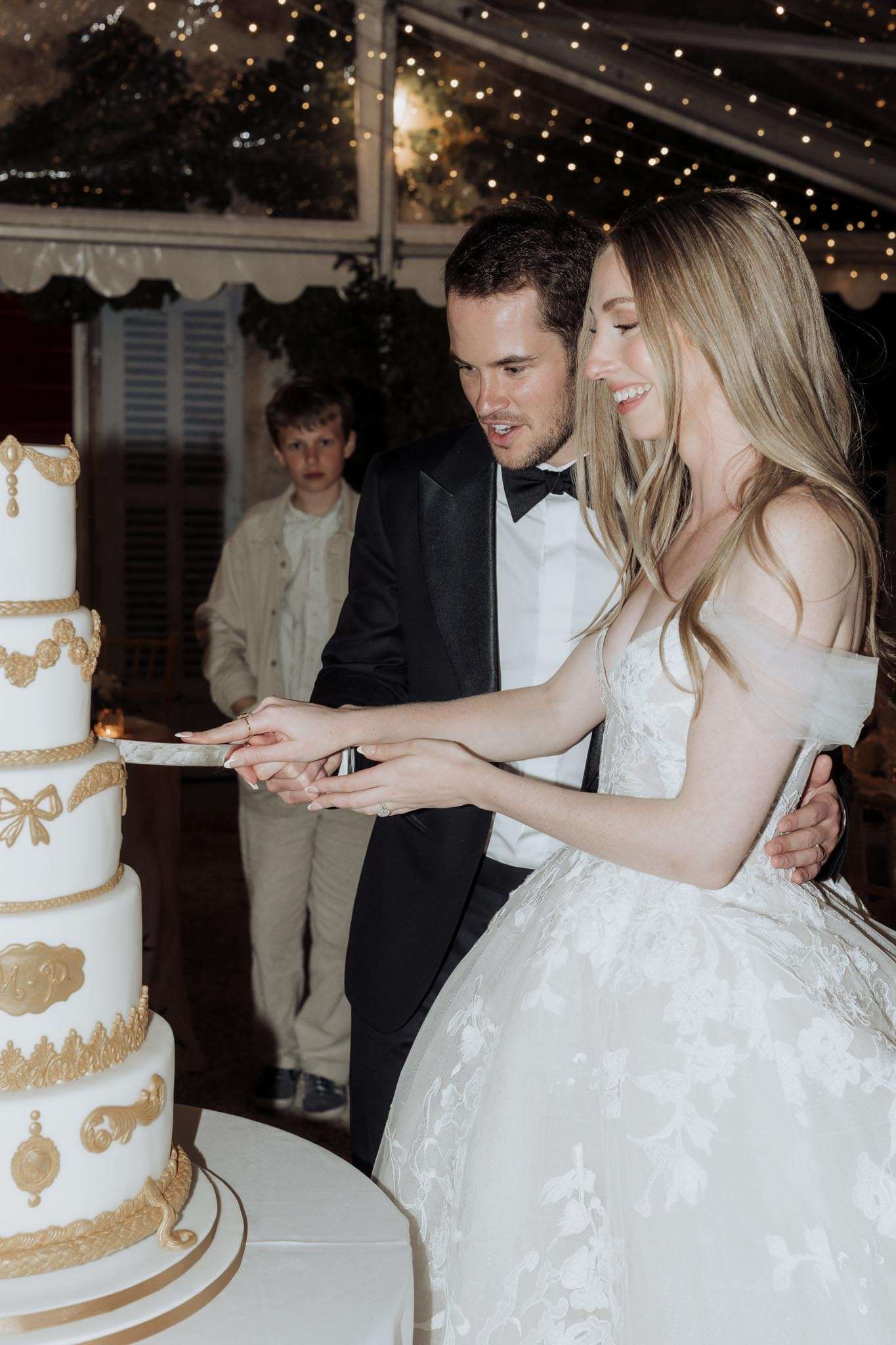 Couple cutting four-tier white wedding cake with gold bas-relief motifs under fairy-lit clear tent