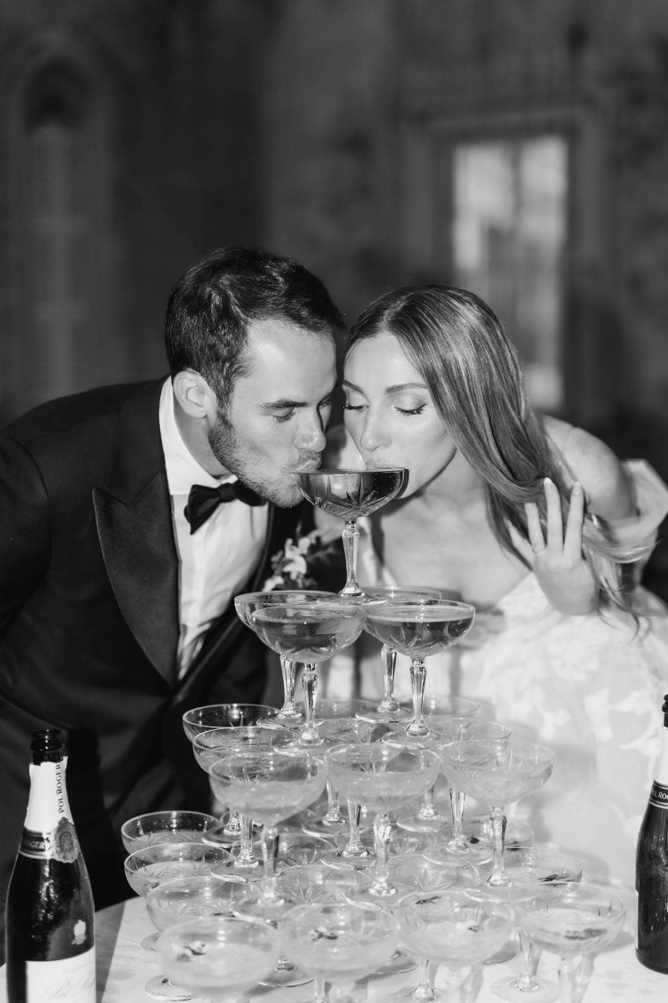 Black and white photo of bride and groom drinking from top of champagne tower with Pol Roger bottles