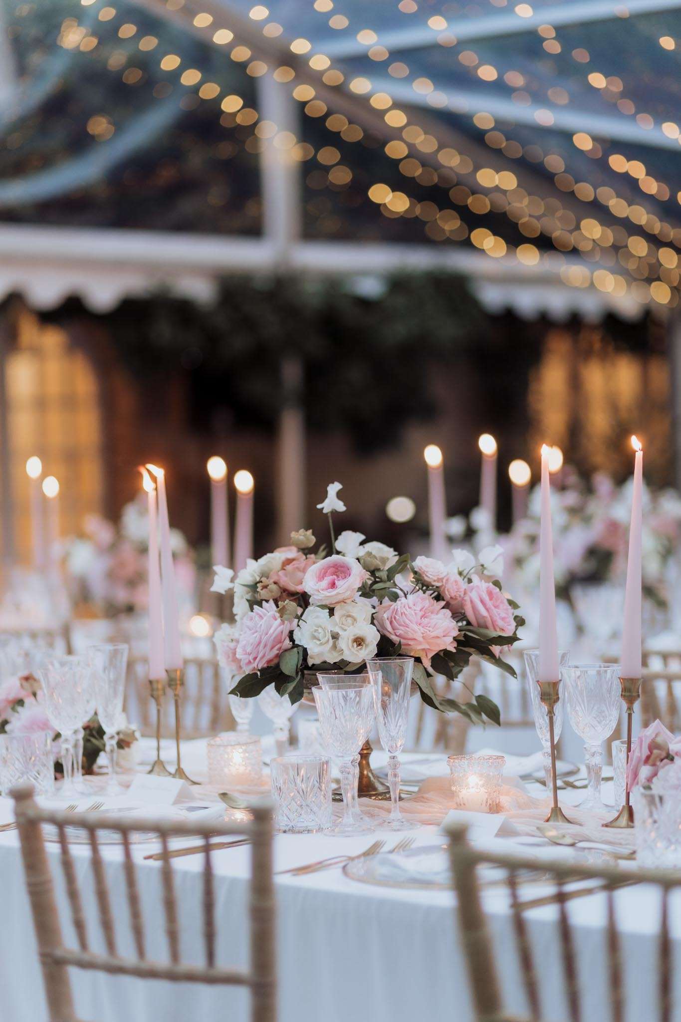 A close-up detail shot of a wedding reception table setting inside a clear-roofed marquee tent strung with warm fairy lights overhead. The round table is dressed in a white linen cloth and set with cut-crystal glassware, gold flatware, and gold chiavari chairs. The centerpiece is a low, lush arrangement of blush pink peonies, pale pink garden roses, ivory spray roses, and dark green foliage. Blush pink taper candles in brass candlestick holders are placed around the centerpiece alongside small votive candles, reinforcing a pink, ivory, and gold color palette. Multiple similarly dressed tables are visible in the soft-focus background, along with a garland-draped structure and warm ambient lighting, indicating a classic romantic reception style.
