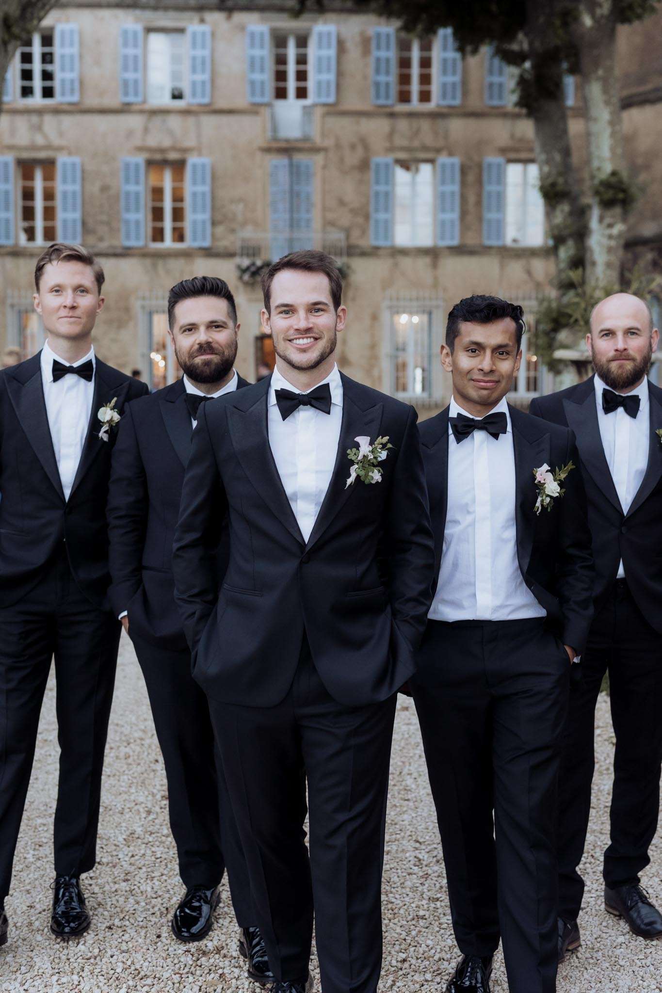 Groom and four groomsmen in black tuxedos walk on gravel driveway in front of French chateau