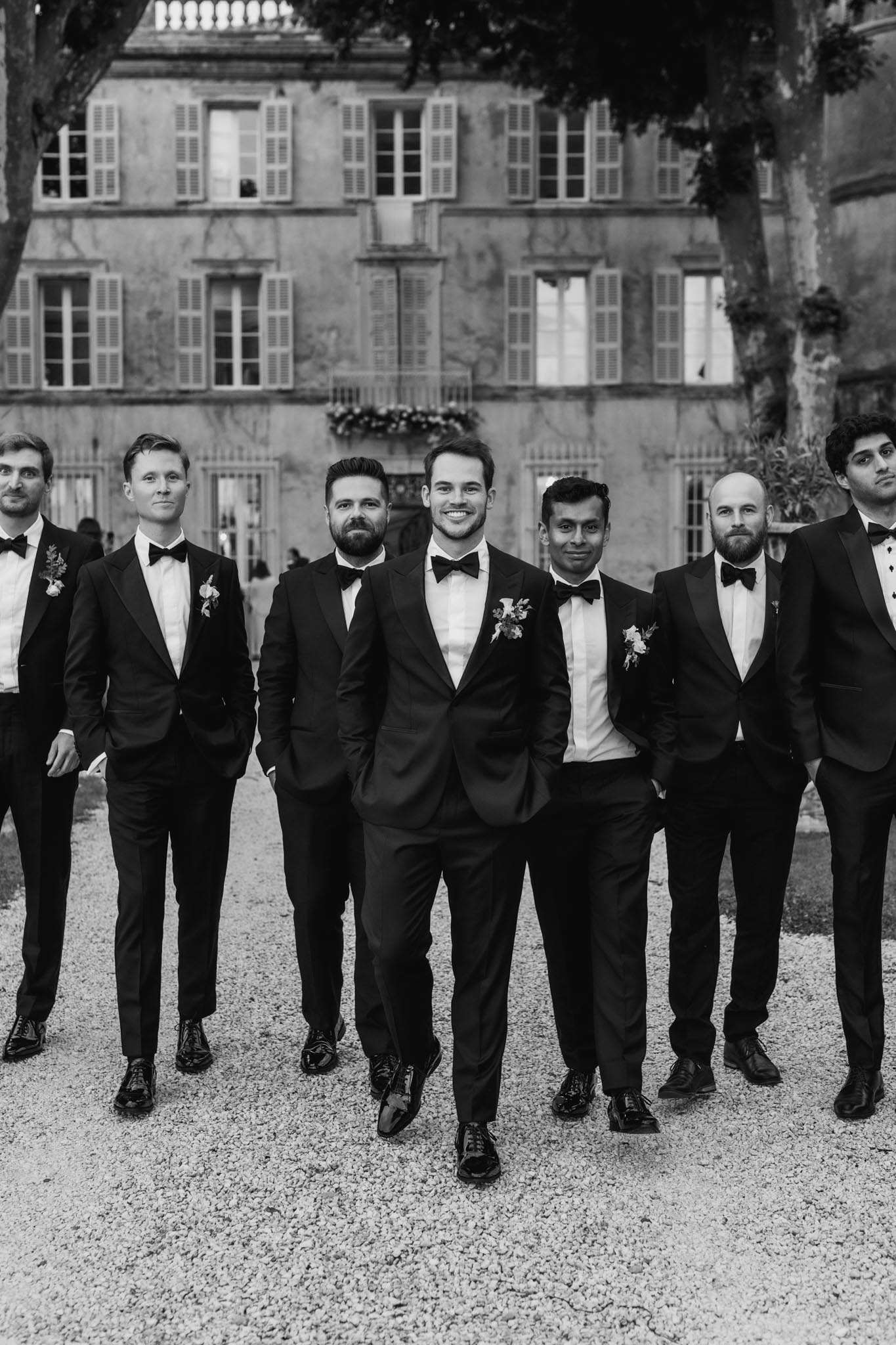 Black and white groom and six groomsmen in tuxedos walking on gravel driveway before shuttered French chateau