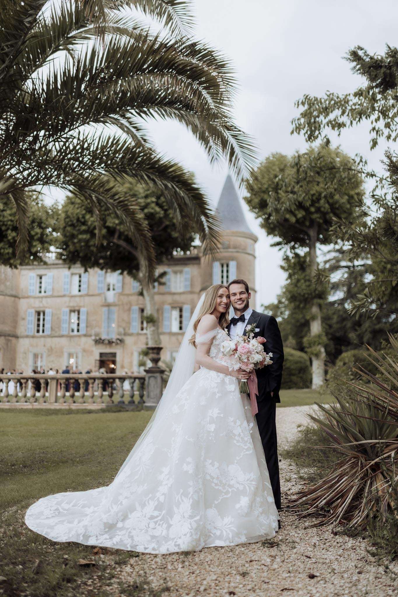 Bride in off-shoulder lace ballgown and groom in navy suit standing on gravel path with chateau in background