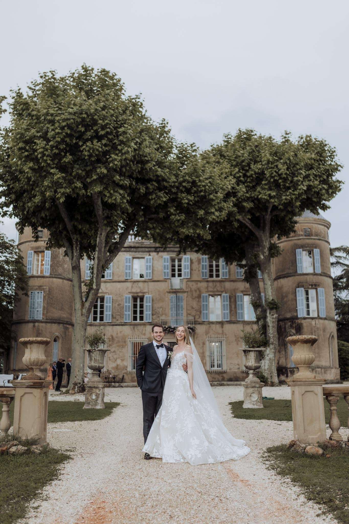 A couple portrait taken outdoors on the gravel driveway of a French château, with the full façade of the property visible behind them. The groom wears a dark navy tuxedo with a black bow tie, and the bride wears a white off-the-shoulder ballgown with floral lace appliqué detailing and a long cathedral-length veil; she holds a small white bouquet. The château features warm ochre stone, pale blue shutters, and rounded tower elements on either side, flanked by large stone urn planters on pedestals. Two guests are faintly visible in the background near the building's entrance. The composition is a full-length portrait shot taken at mid-distance, centering the couple symmetrically within the driveway. Potential venue feature image.