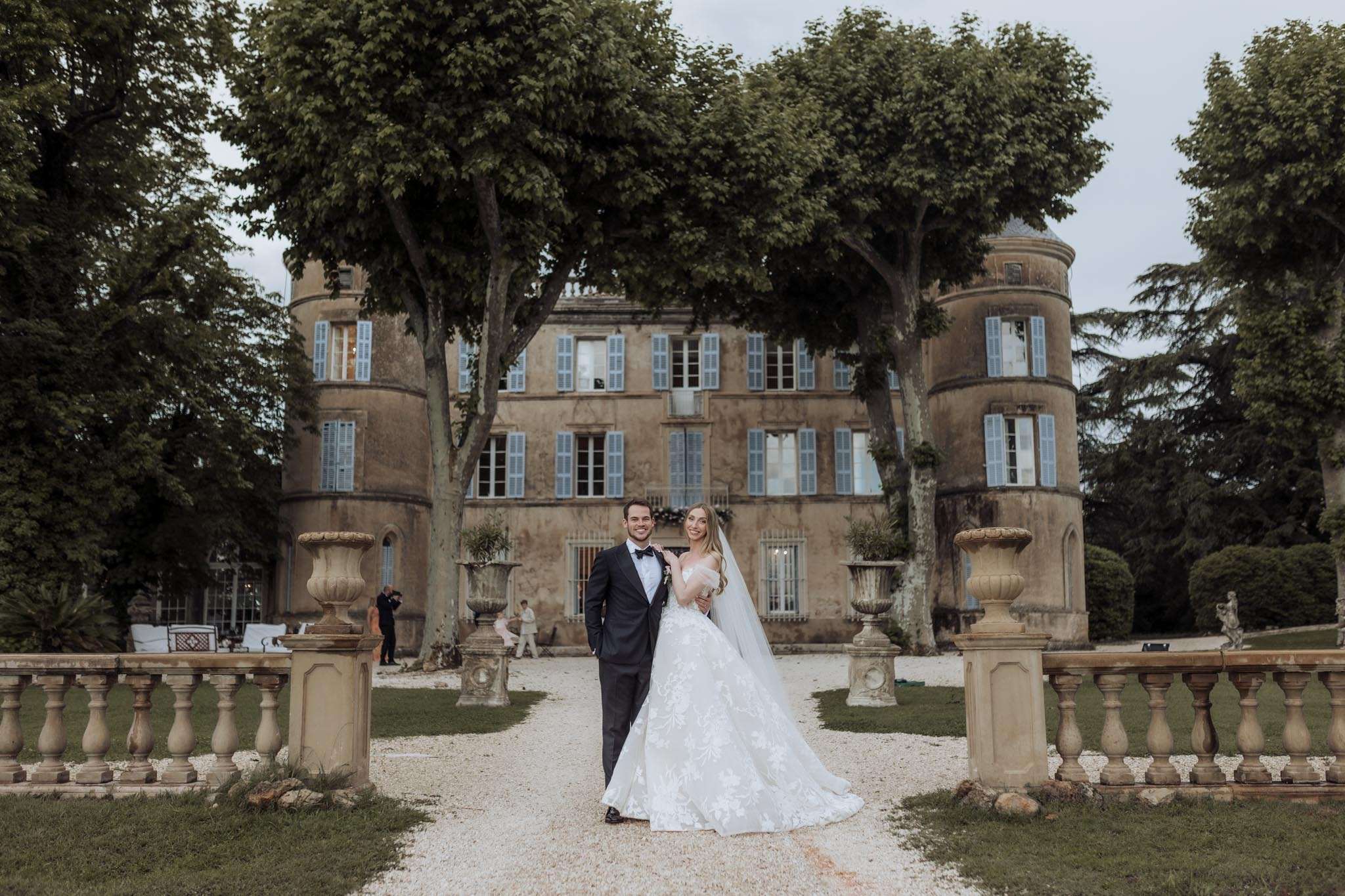 A couple poses for a portrait on the gravel driveway leading up to a French château, flanked by stone balustrade terracing and large decorative urns on pedestals. The groom wears a dark navy tuxedo with a black bow tie and a white boutonniere, while the bride wears a full ballgown with an ivory lace floral appliqué skirt and a long cathedral-length veil, the dress featuring an off-the-shoulder or strapless silhouette. The château behind them is a multi-story ochre stone building with pale blue shutters and two rounded corner towers, with a photographer and at least one other person visible in the background near the entrance. The composition is a wide portrait shot that equally showcases the couple and the full façade of the venue. Potential venue feature image.