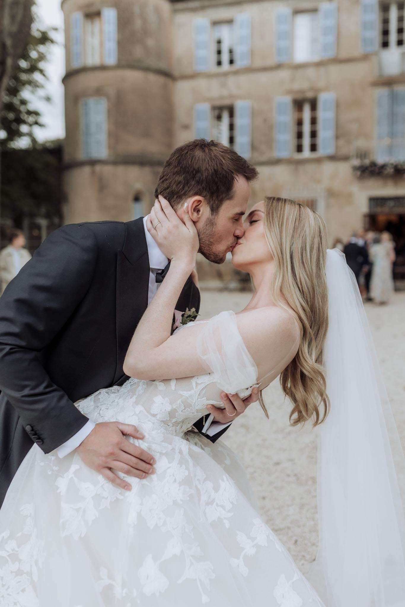 A couple shares a kiss in the courtyard of a French château, captured in a close-up portrait shot with the building softly blurred in the background. The bride wears a white ballgown with floral lace appliqué throughout the skirt and sheer off-the-shoulder puff sleeves, paired with a long flowing veil and loose wavy blonde hair. The groom is dressed in a dark charcoal tuxedo with a bow tie and a small floral boutonnière featuring pale pink blooms. Several guests are visible in the blurred background near the château entrance, which features pale stone architecture with light blue shutters and a circular tower.